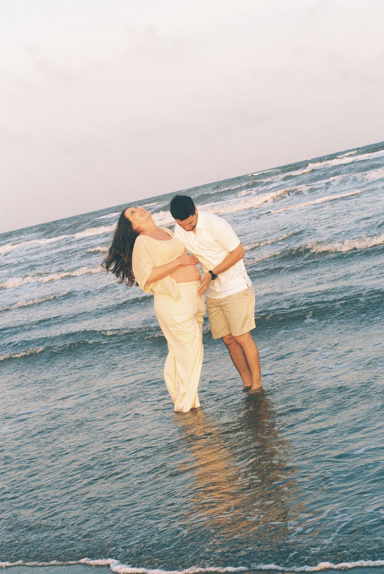 Couple playing in ocean waves during sunset beach maternity photoshoot.