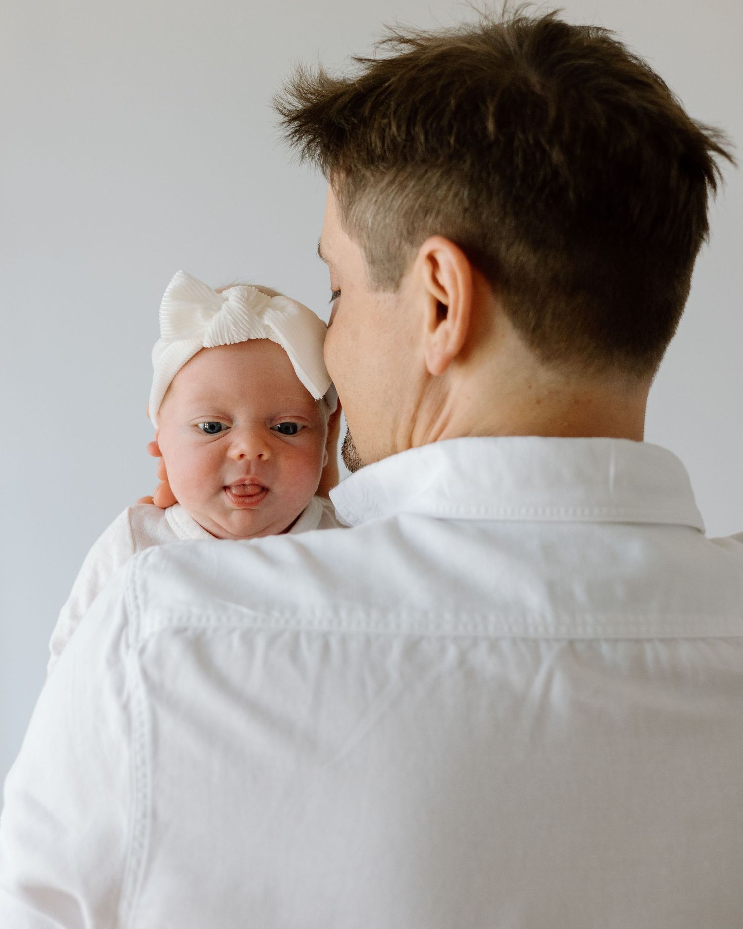 Baby wearing white bow headband being held by parent in white shirt.