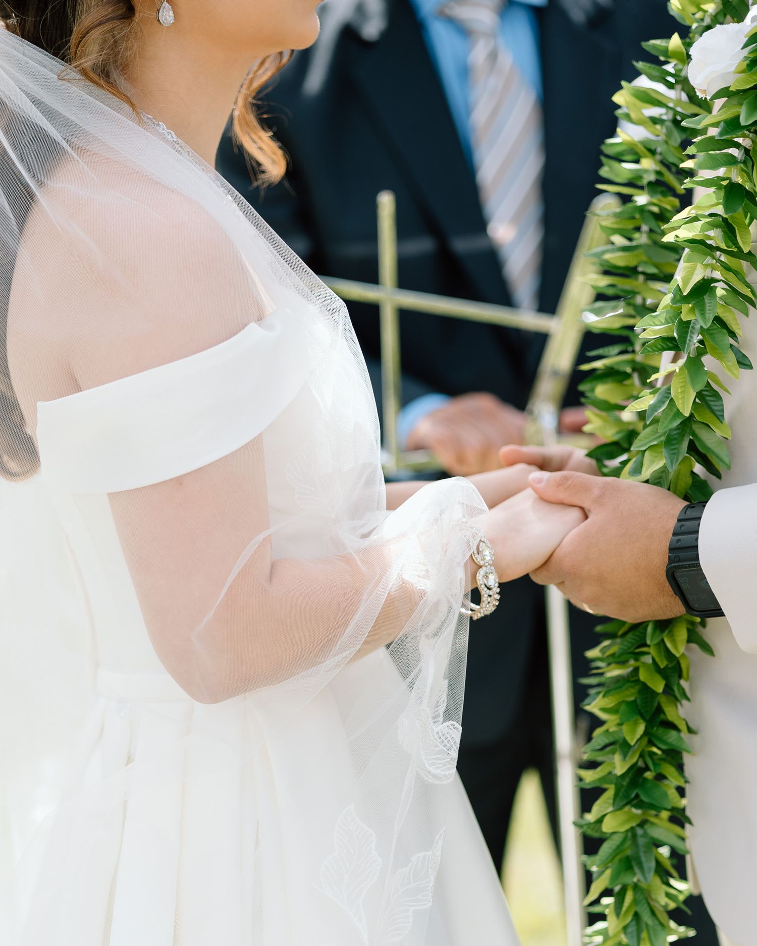 Close-up of wedding ring exchange with traditional white bridal gown and green lei detail.