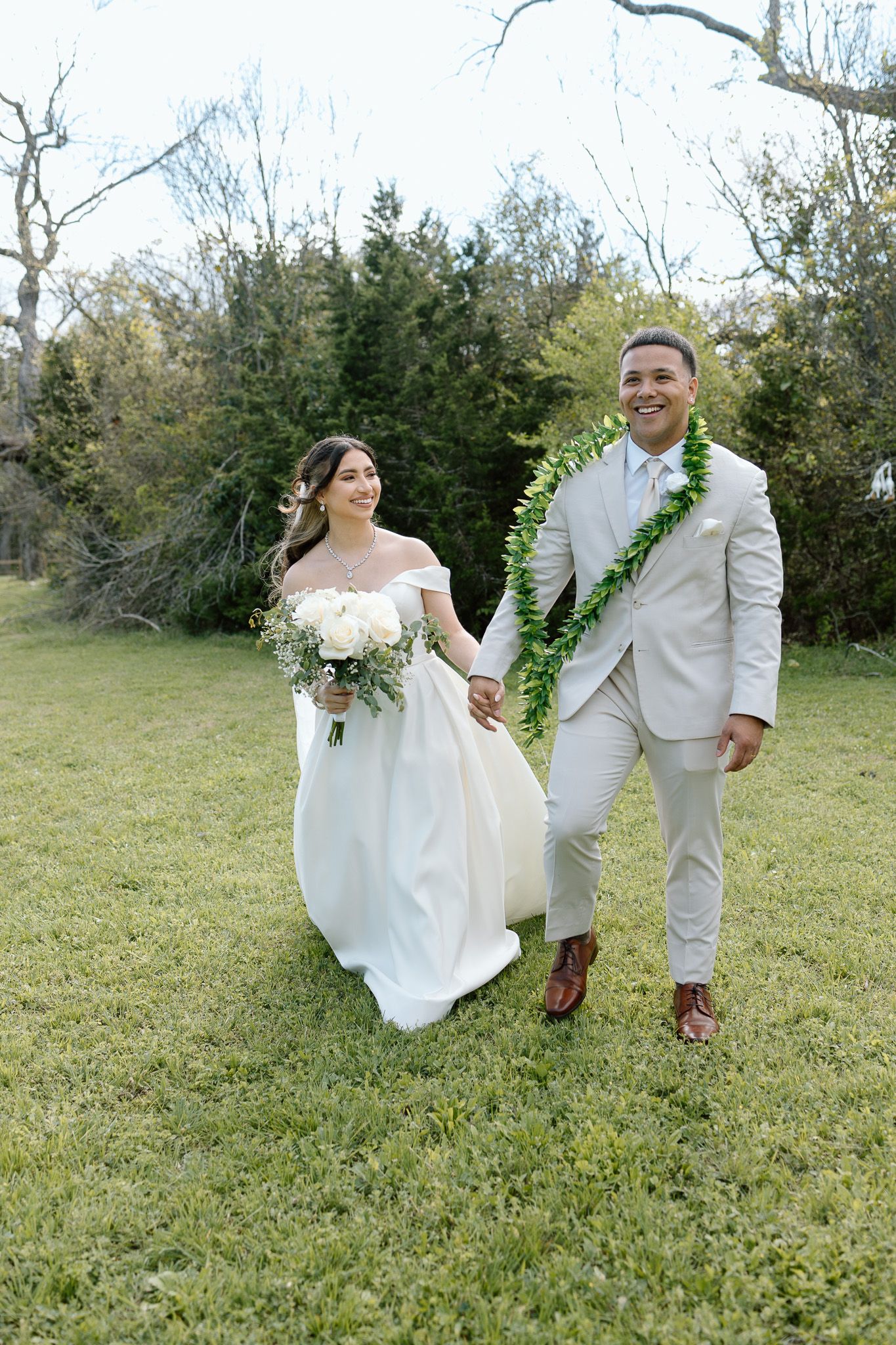 Newlyweds walking hand in hand on grass, groom wearing traditional Hawaiian green lei.