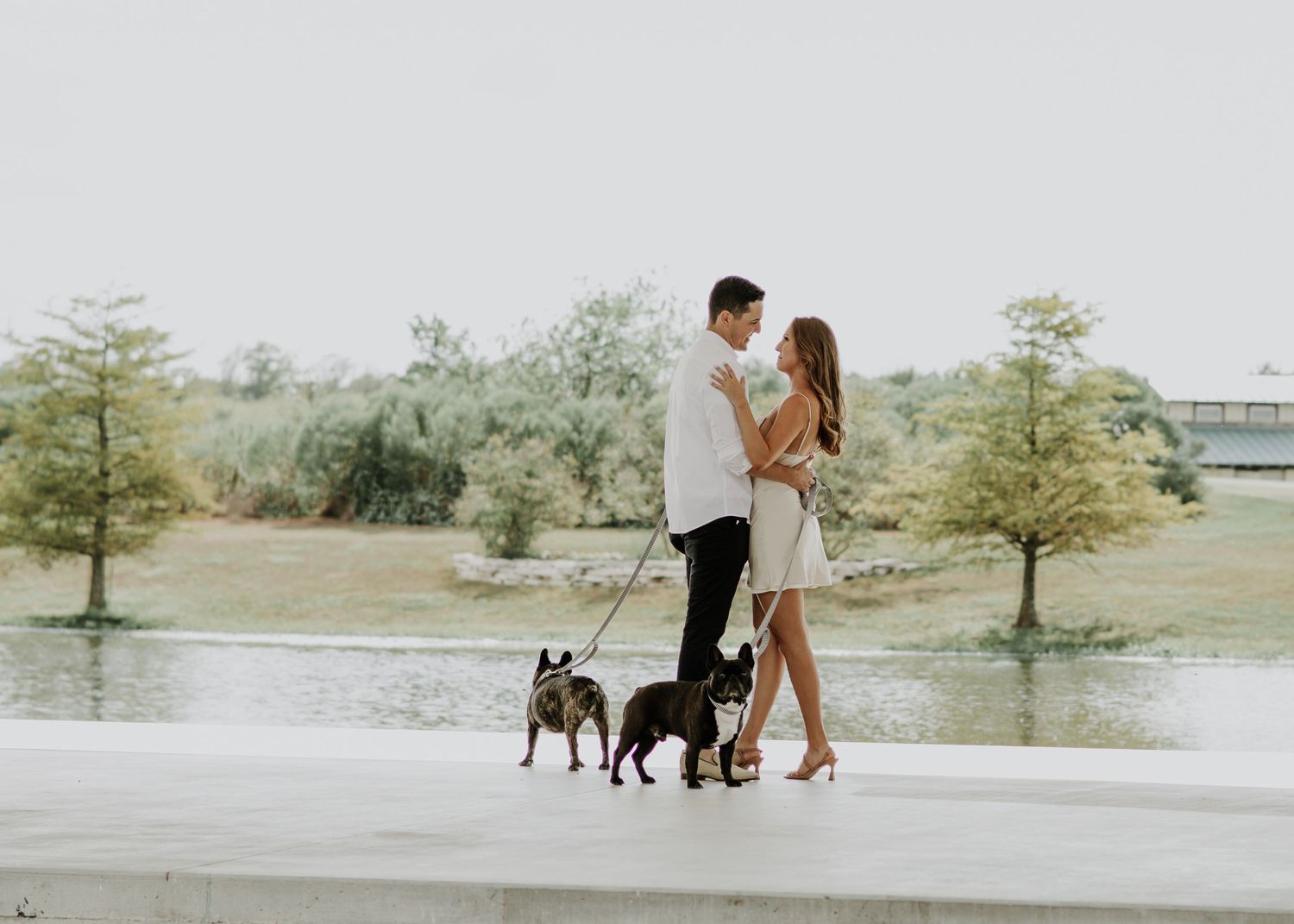 A couple stands close together on a walkway by a lake with two small dogs.