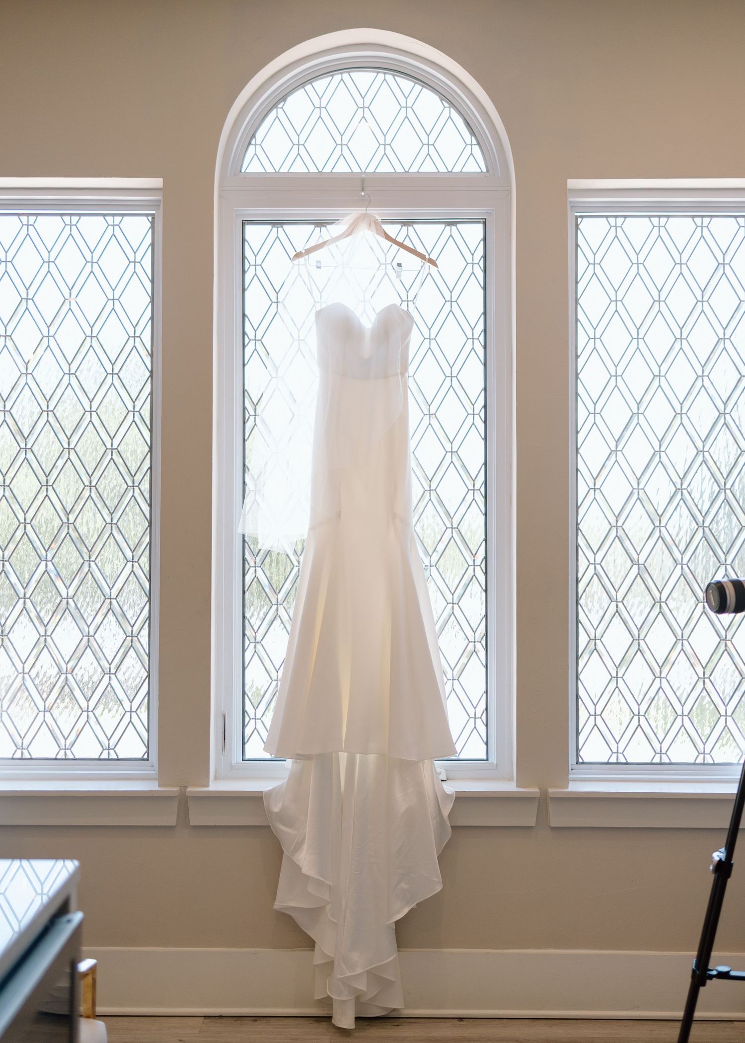Elegant white wedding dress hanging in front of arched window with diamond pattern glass.