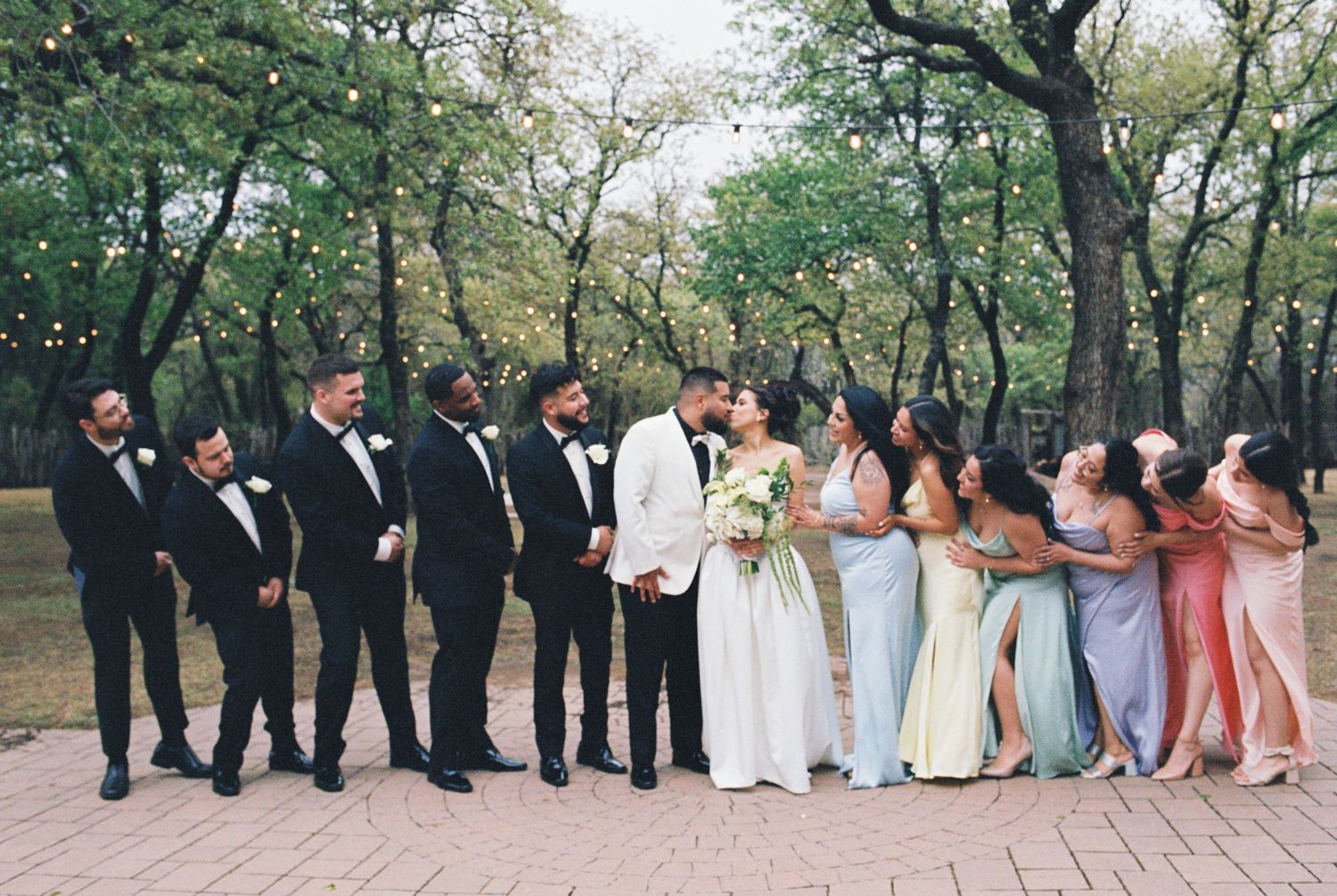 Wedding party poses together with bridesmaids in pastel gowns and groomsmen in black suits.