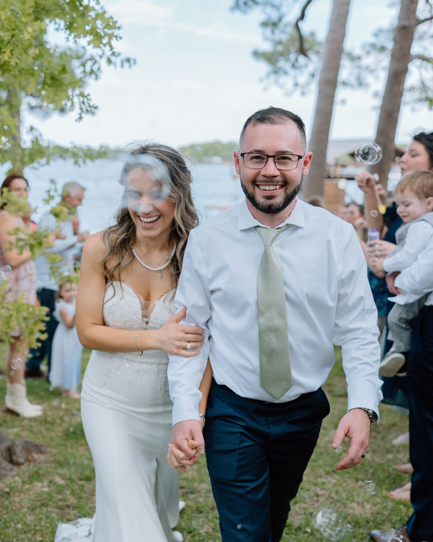 Joyful newlyweds walk hand in hand during outdoor lakeside celebration.