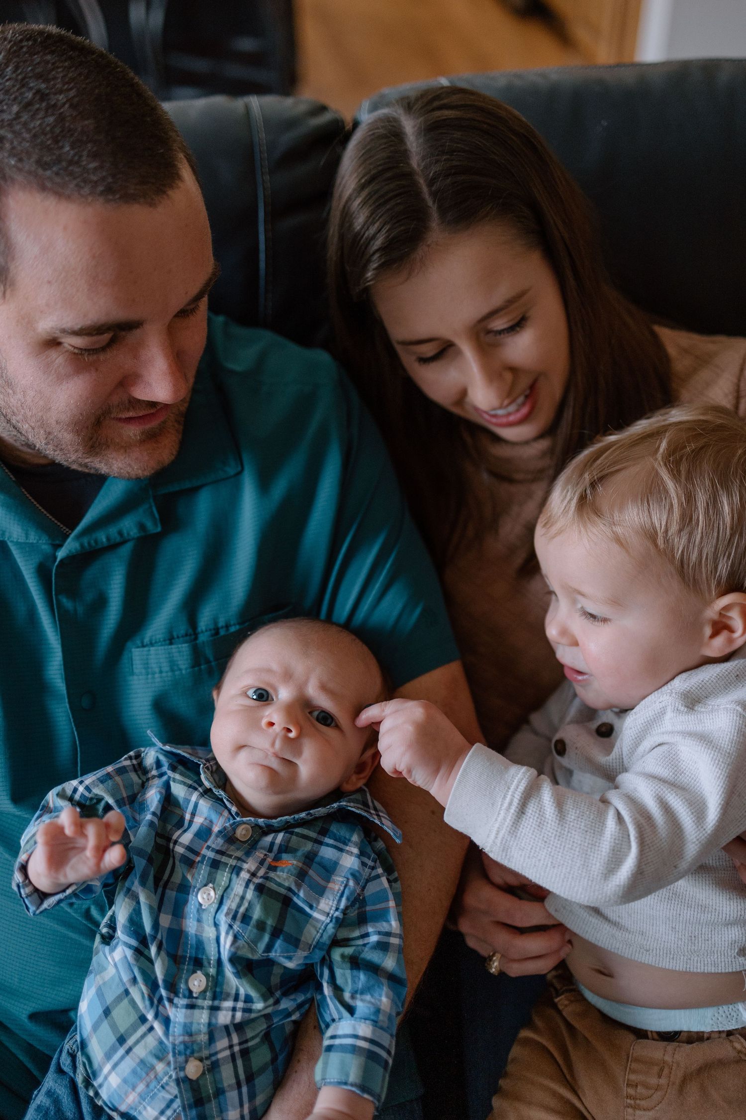 A young toddler gently touches a baby sibling while parents look on lovingly from their cozy couch.