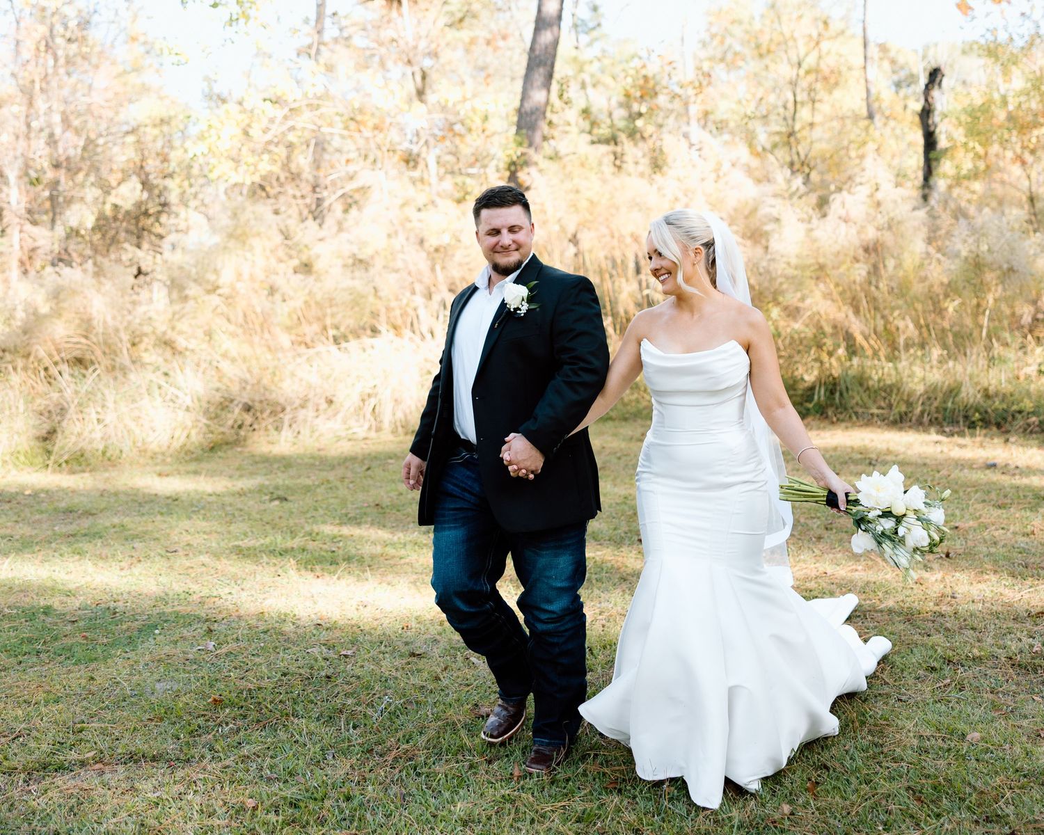 Newlywed couple walks hand in hand through golden autumn field, bride holding white bouquet.