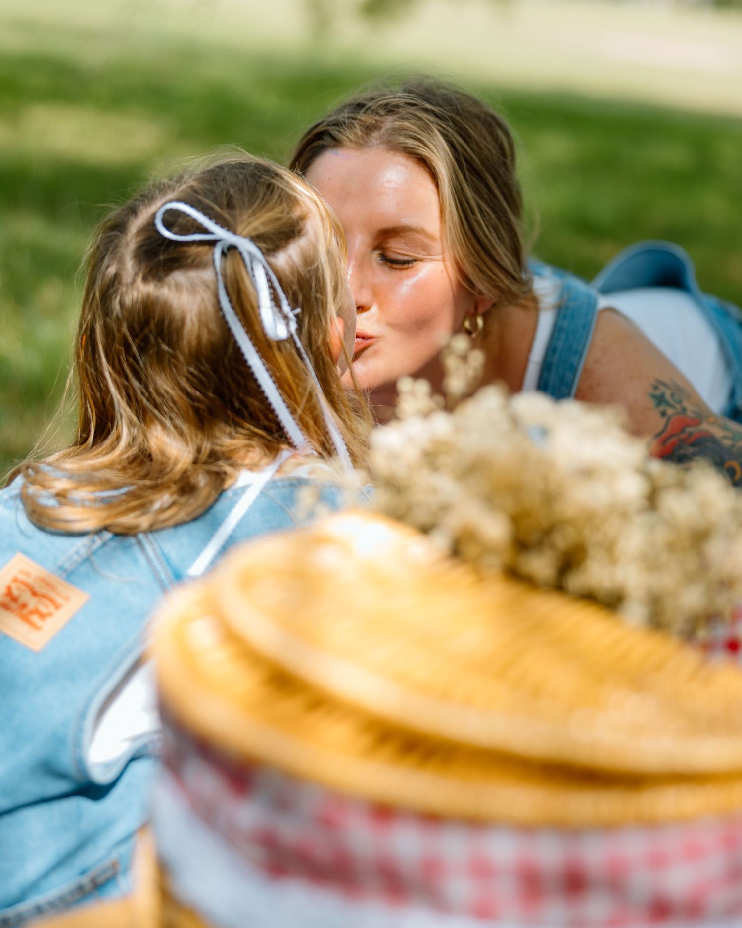 A mother kisses her young daughter during a sunny outdoor picnic, with a wicker basket and wildflowers in the foreground.