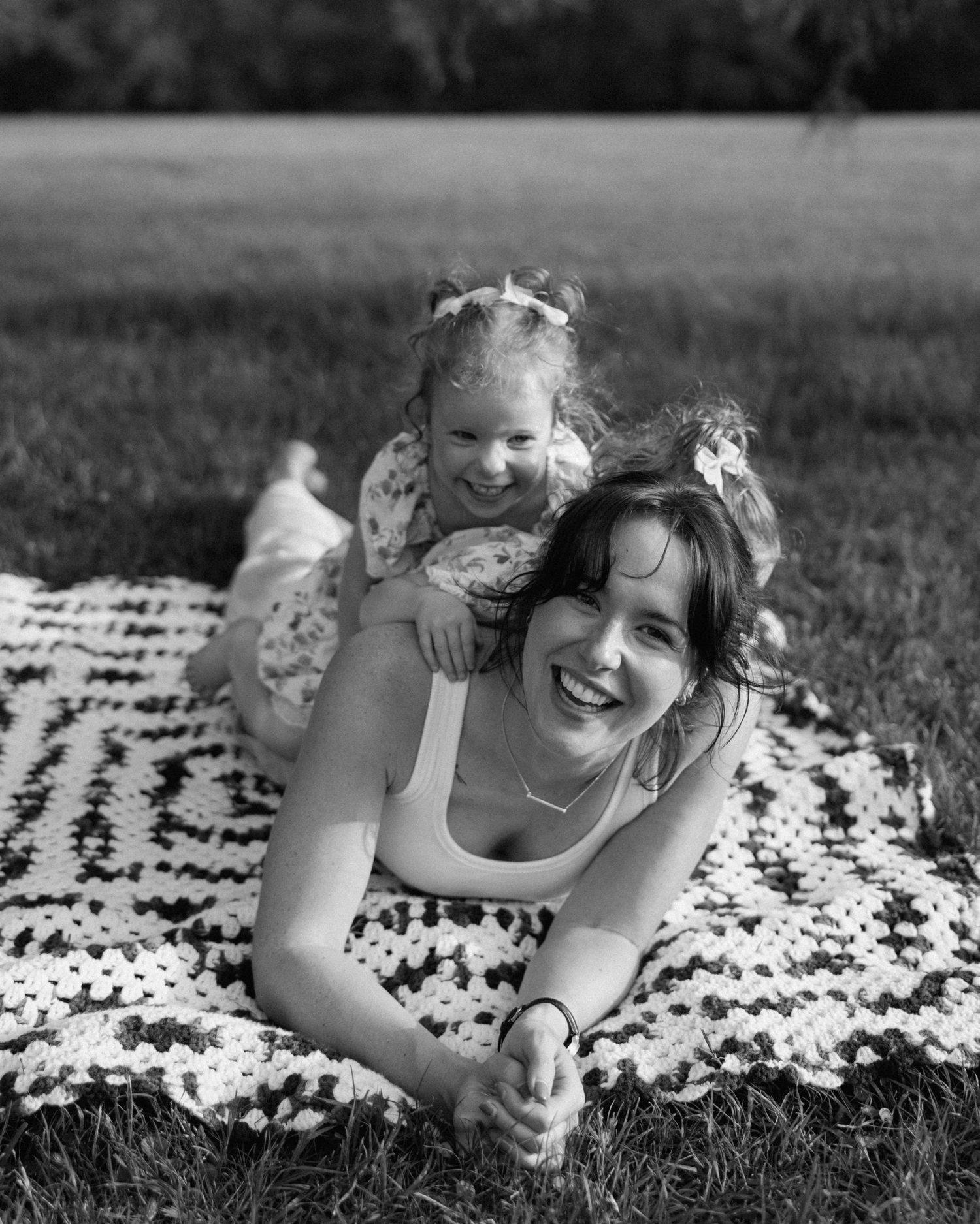 Black and white photo of a laughing woman lying on a blanket outdoors with a smiling toddler girl on her back.