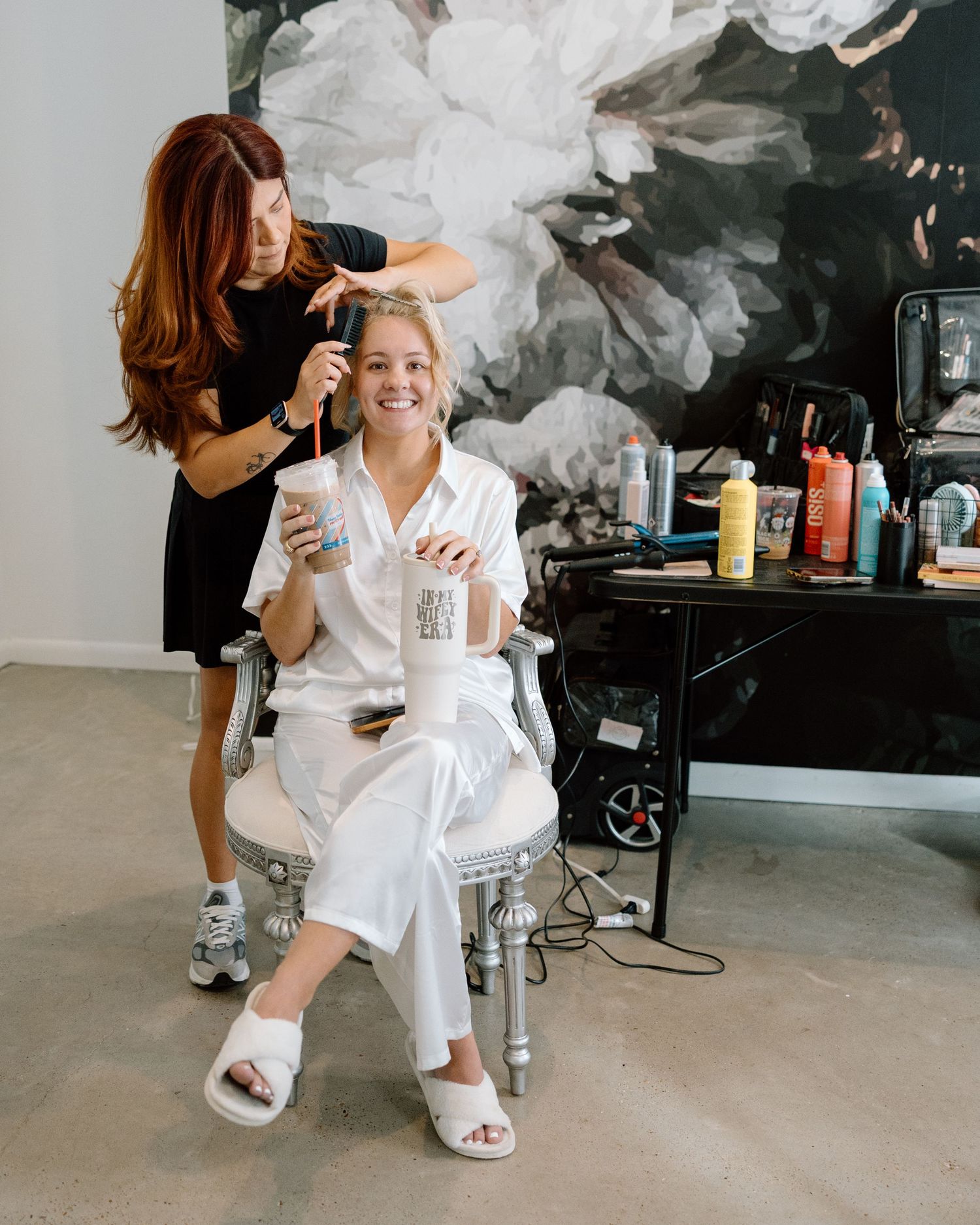 Bride smiling in white robe holding a drink while hairstylist works on her hair in a modern salon.