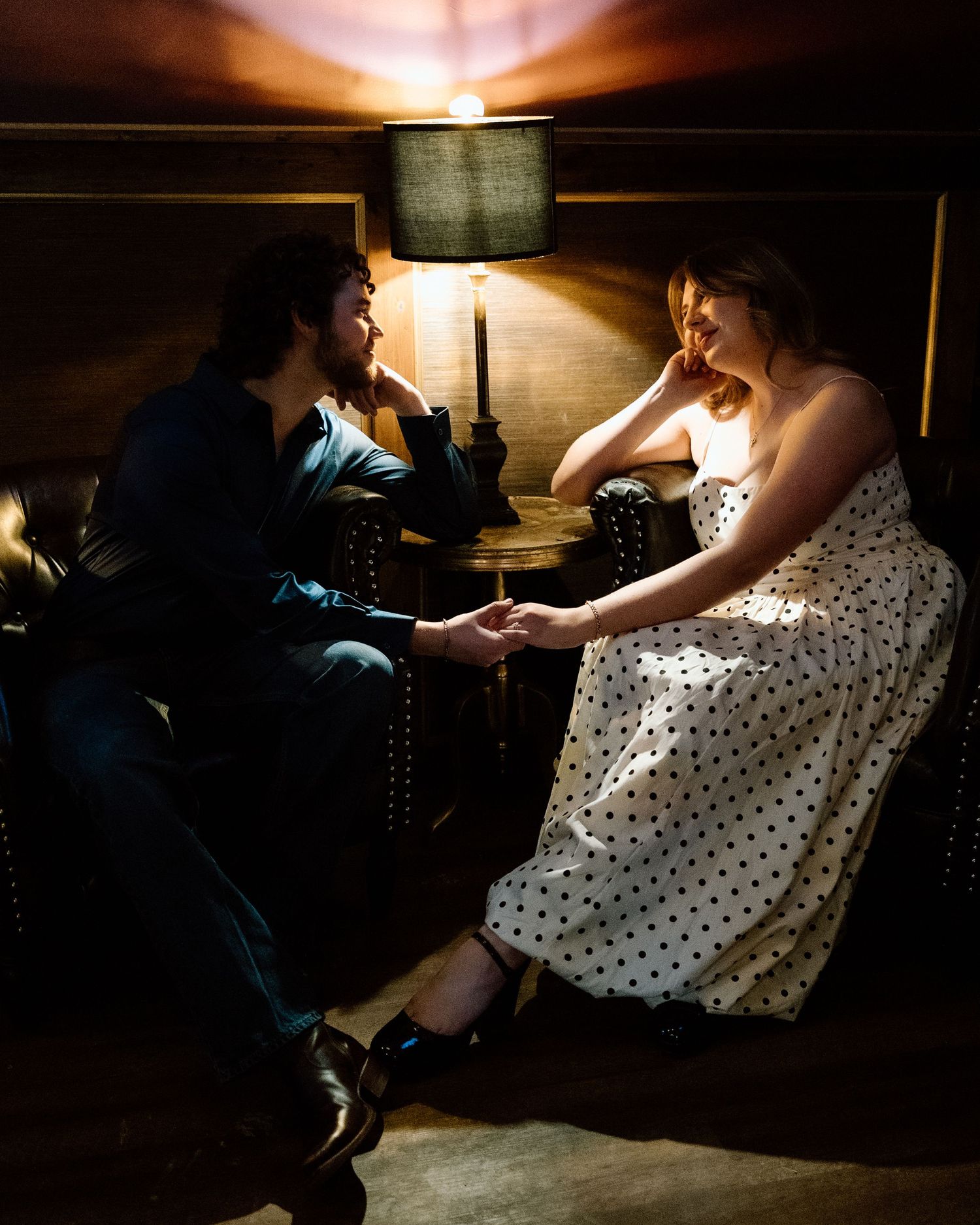 A couple holds hands in a dimly lit room, the woman in a polka dot dress and man in dark attire sharing an intimate moment.