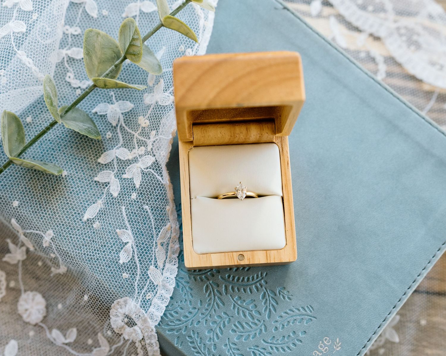 A delicate solitaire diamond engagement ring displayed in an open wooden ring box on a blue lace fabric.