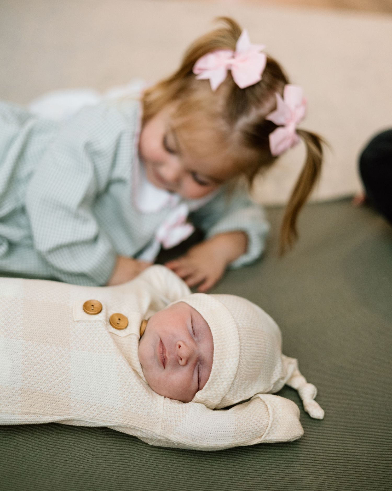 Toddler girl with pink bows leaning over to look at sleeping swaddled newborn baby on green carpet.