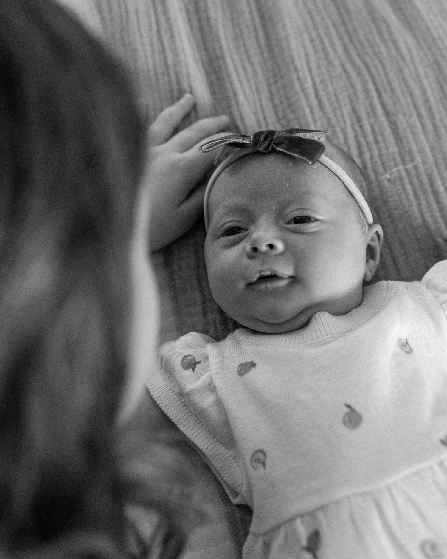 Black and white photo of a newborn baby girl wearing a bow headband, looking up at a person touching her head.