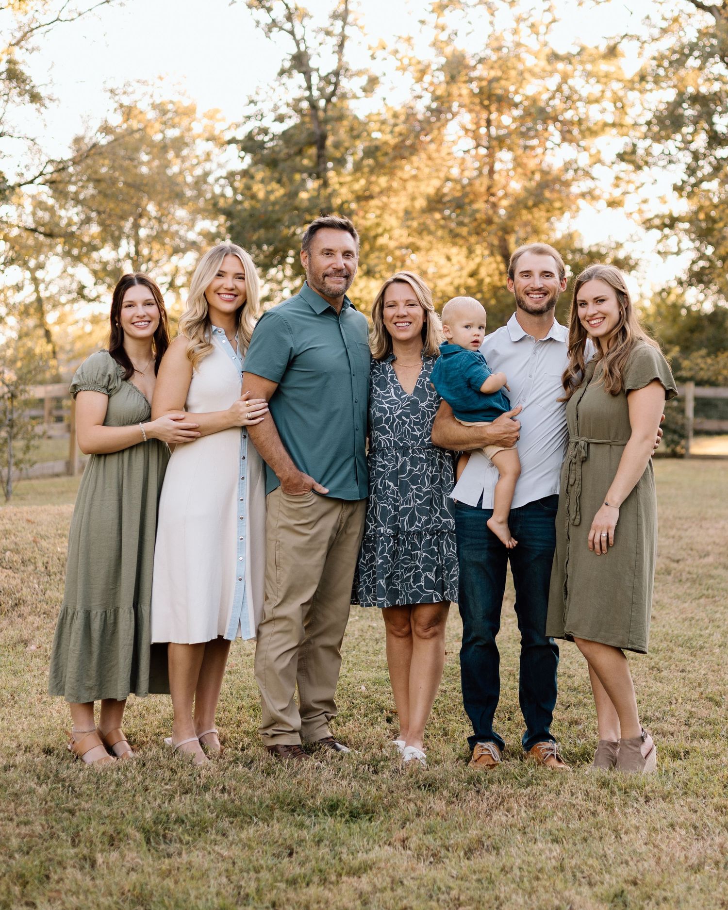 A joyful family of seven poses outdoors at golden hour, including a baby, in coordinated earth-tone outfits.