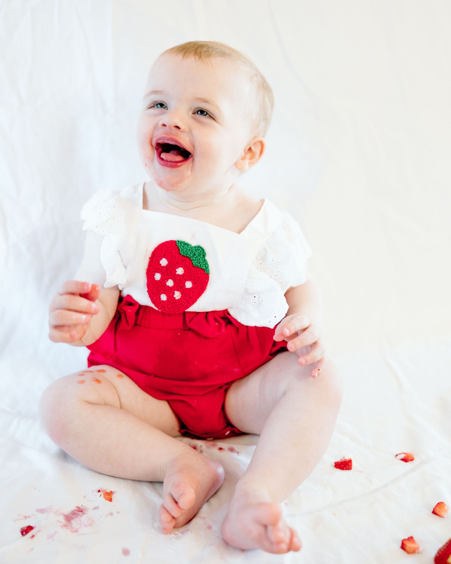 Happy baby girl laughing in red and white strawberry romper during first birthday cake smash session.