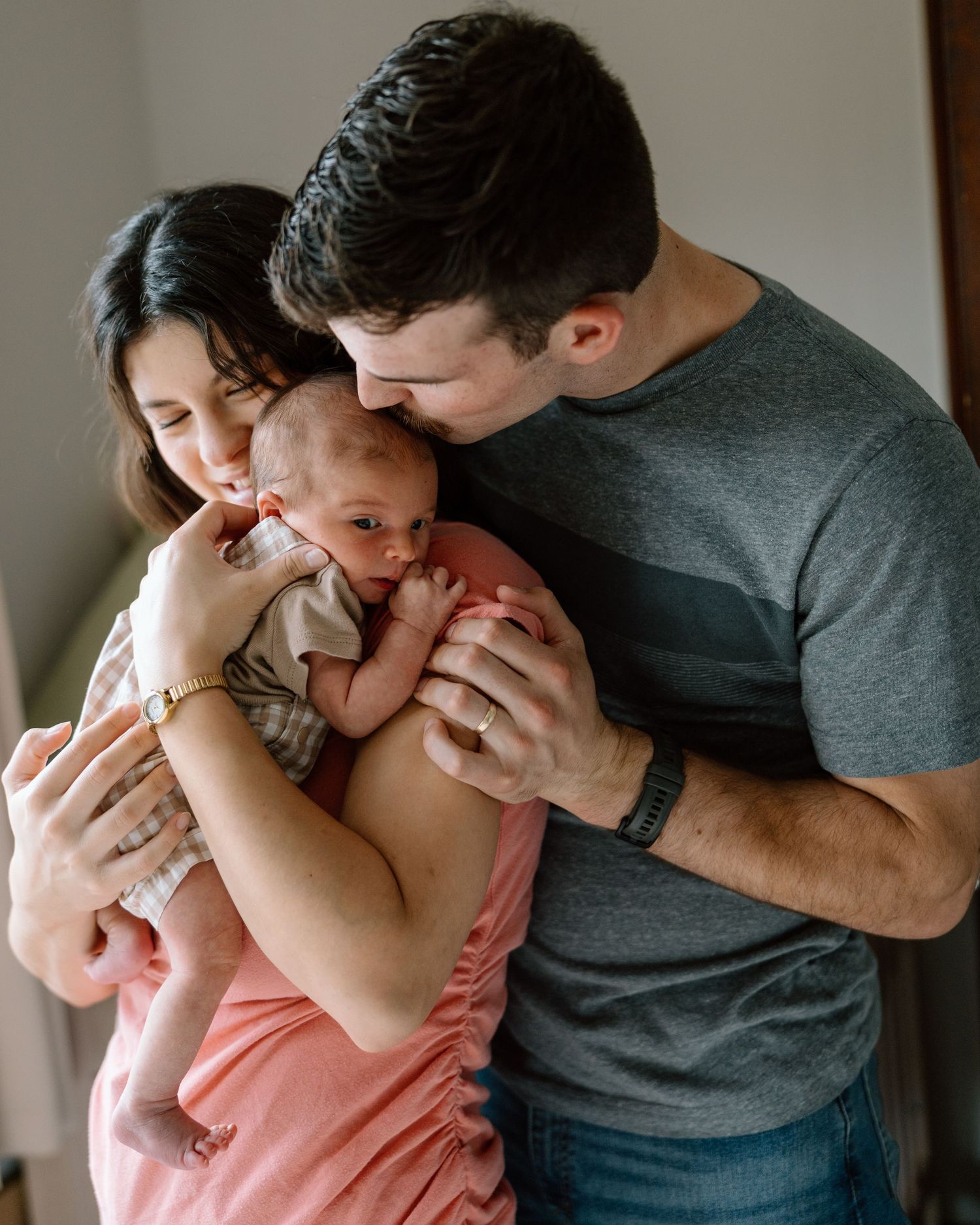 A loving couple shares a tender moment kissing their newborn baby, with the mother in a pink dress.