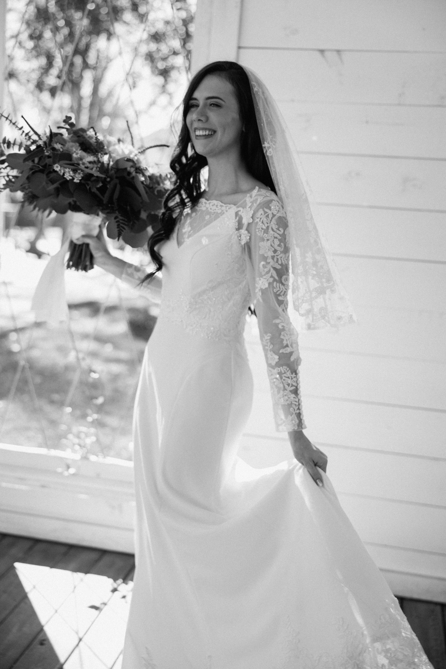 Black and white photo of bride in lace gown holding floral bouquet with veil.