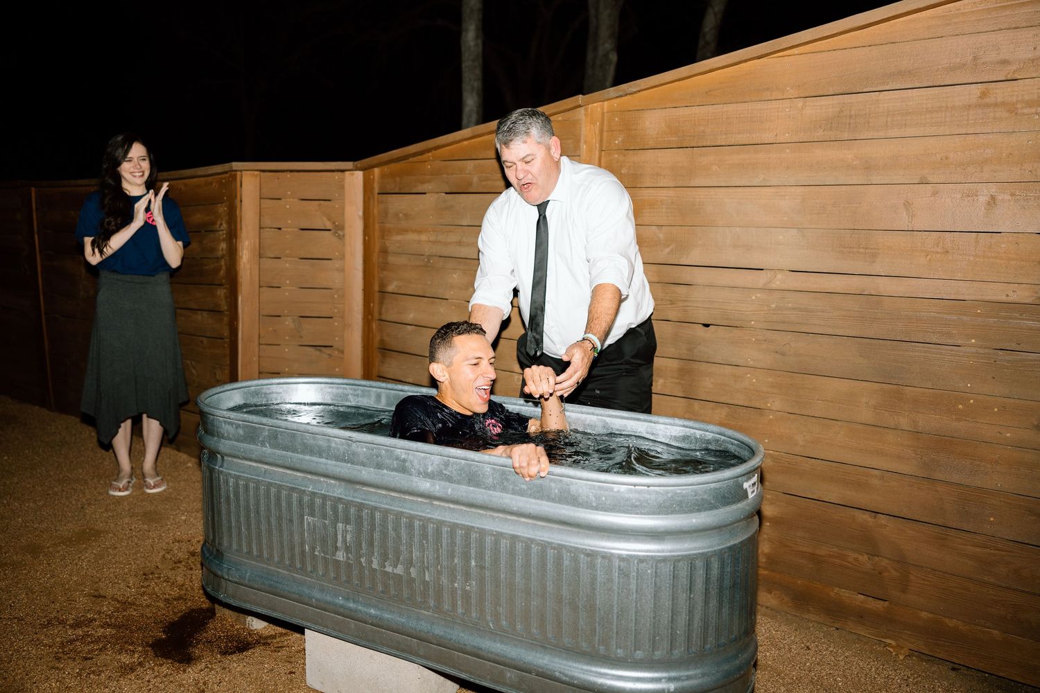 Man assisting young boy during outdoor baptism in galvanized metal trough at nighttime ceremony.