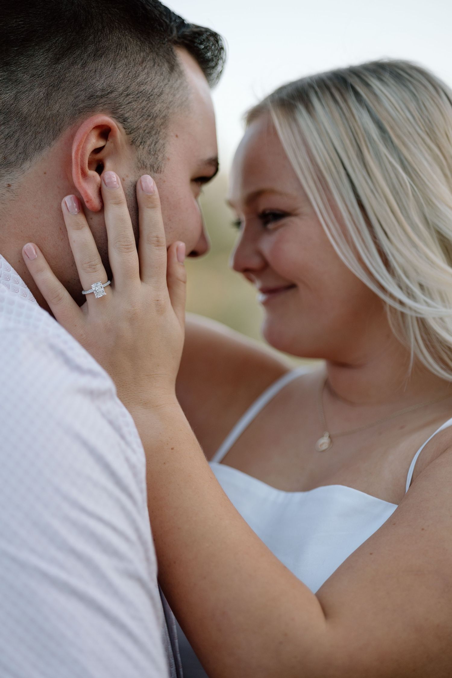 Couple sharing a romantic moment, woman with engagement ring touching man's face, smiling closely outdoors.