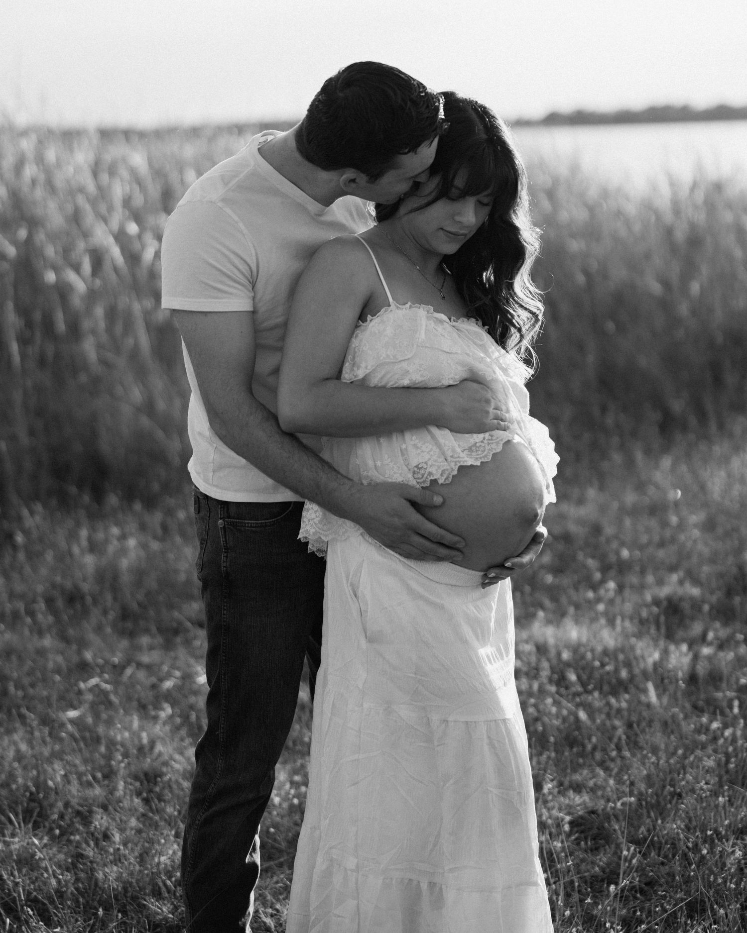 Black and white maternity photo of couple embracing outdoors, man kissing womans head while holding her pregnant belly.