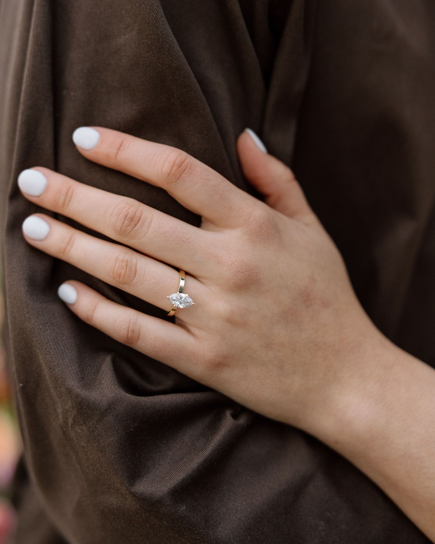 A delicate gold engagement ring with a marquise diamond sits on a woman's finger with white nail polish.