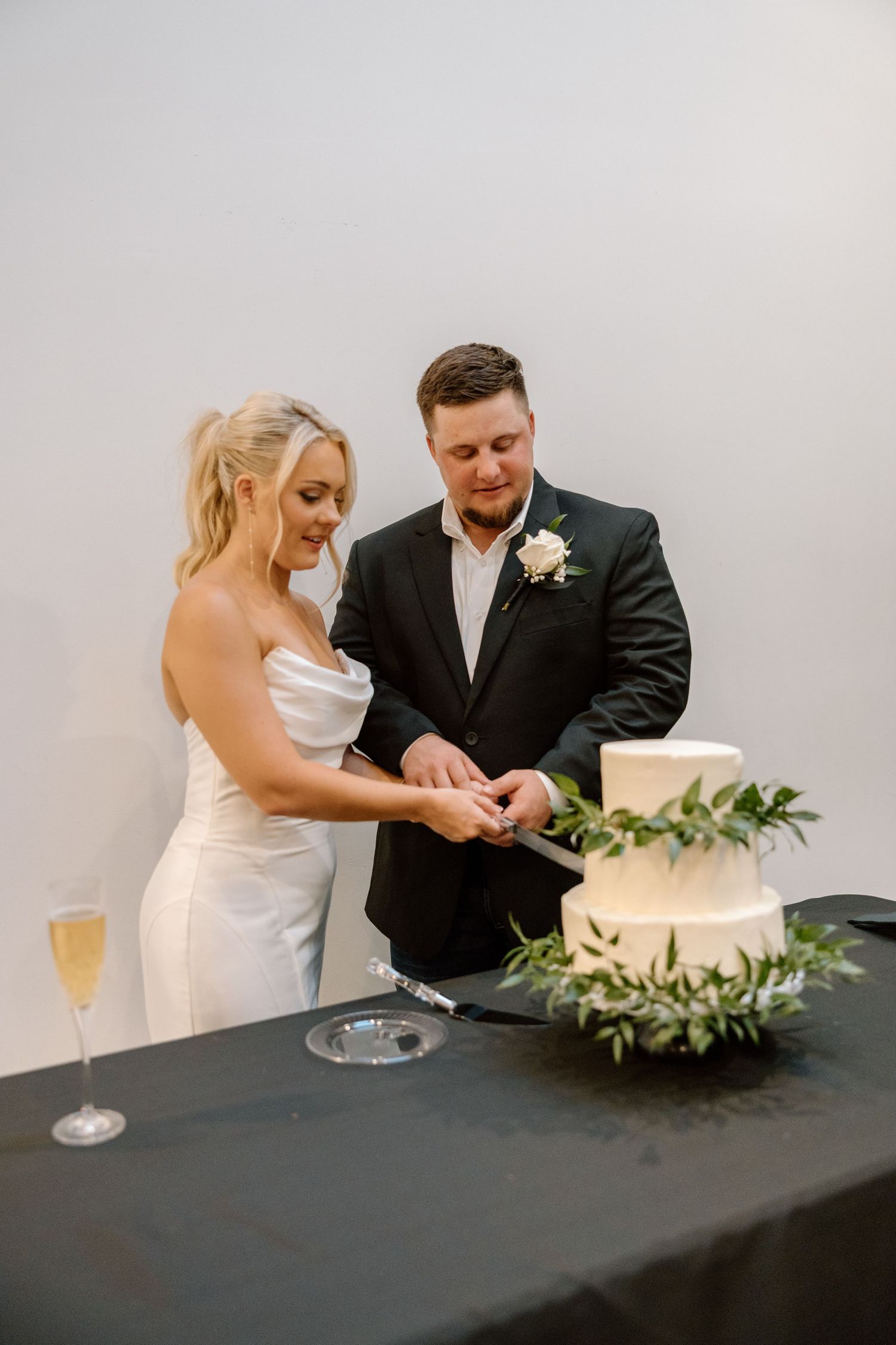 Bride and groom cut elegant two-tier white wedding cake decorated with greenery at reception table.
