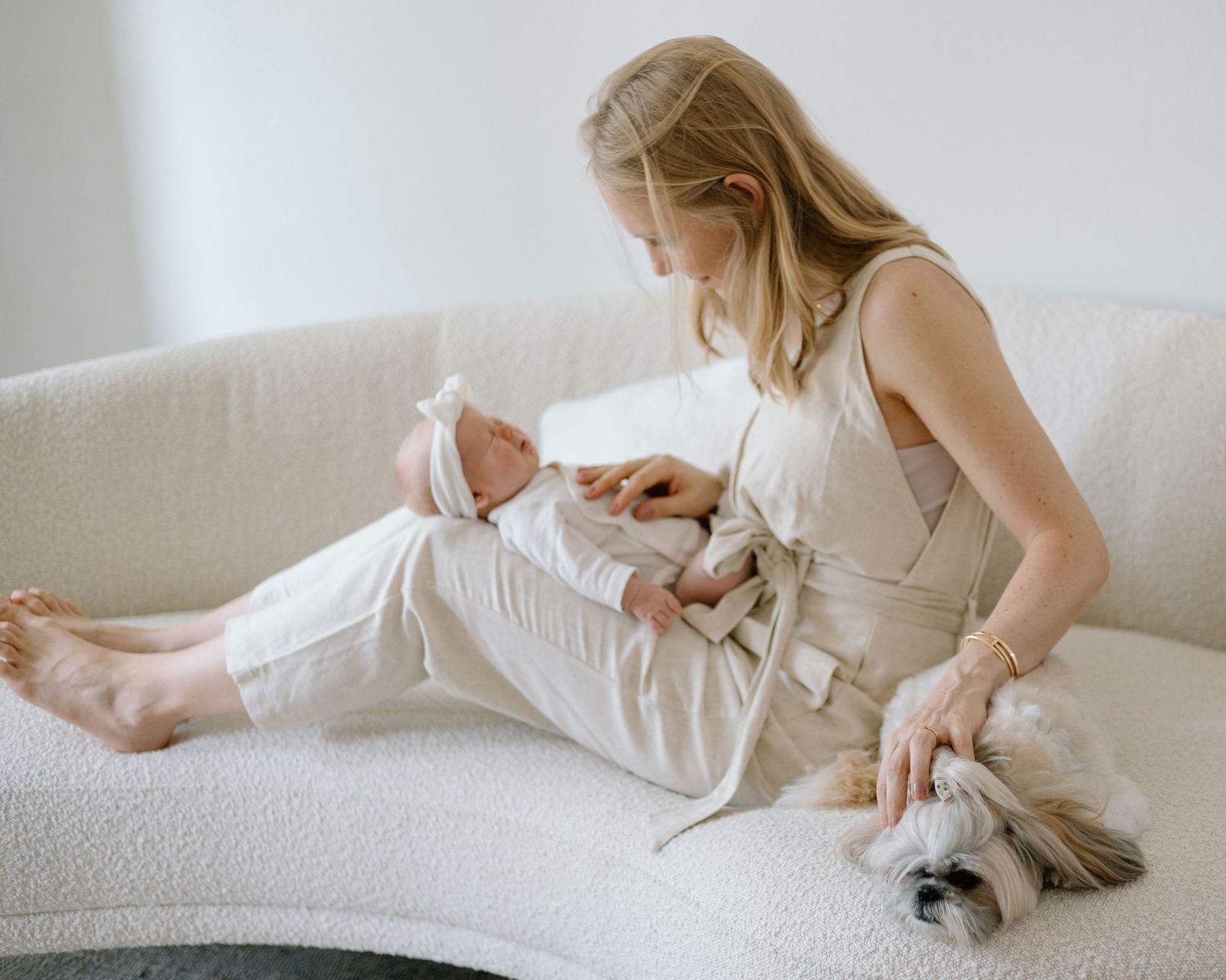 Woman in white dress sitting on couch with baby while small dog rests nearby.