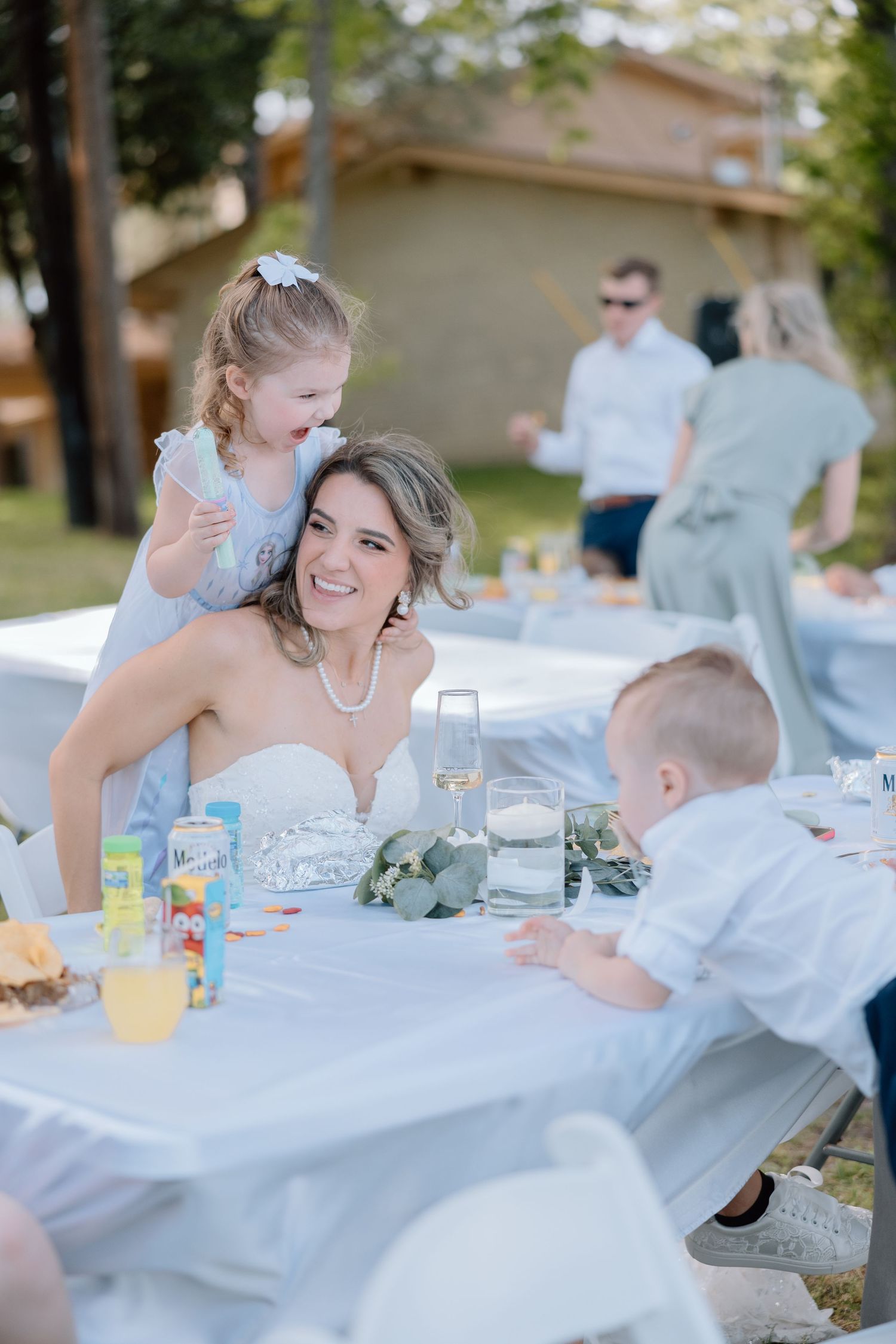 An outdoor wedding reception with guests gathered around a white-clothed table.