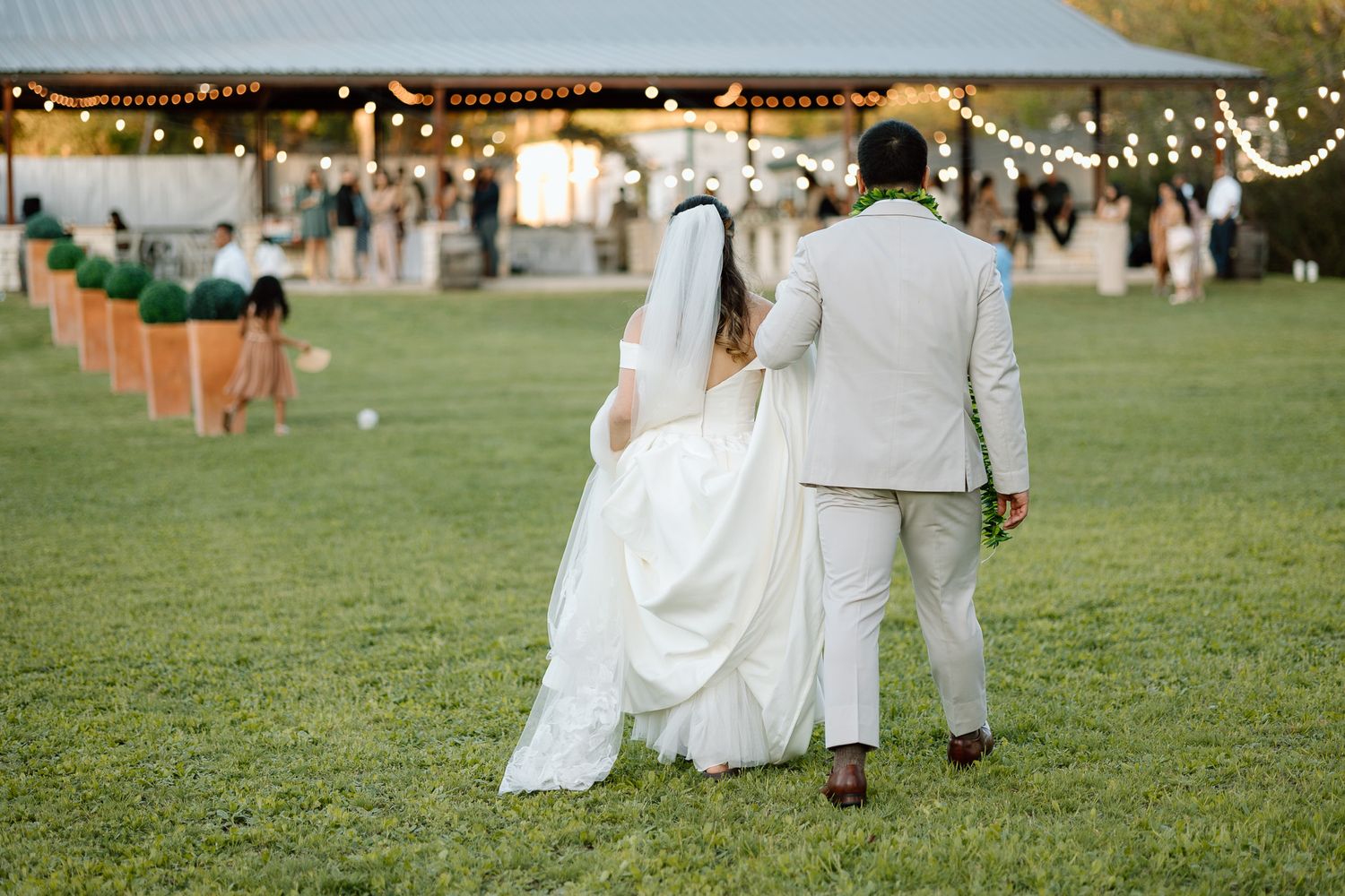 Newlyweds walking away from ceremony site with string lights overhead at dusk.