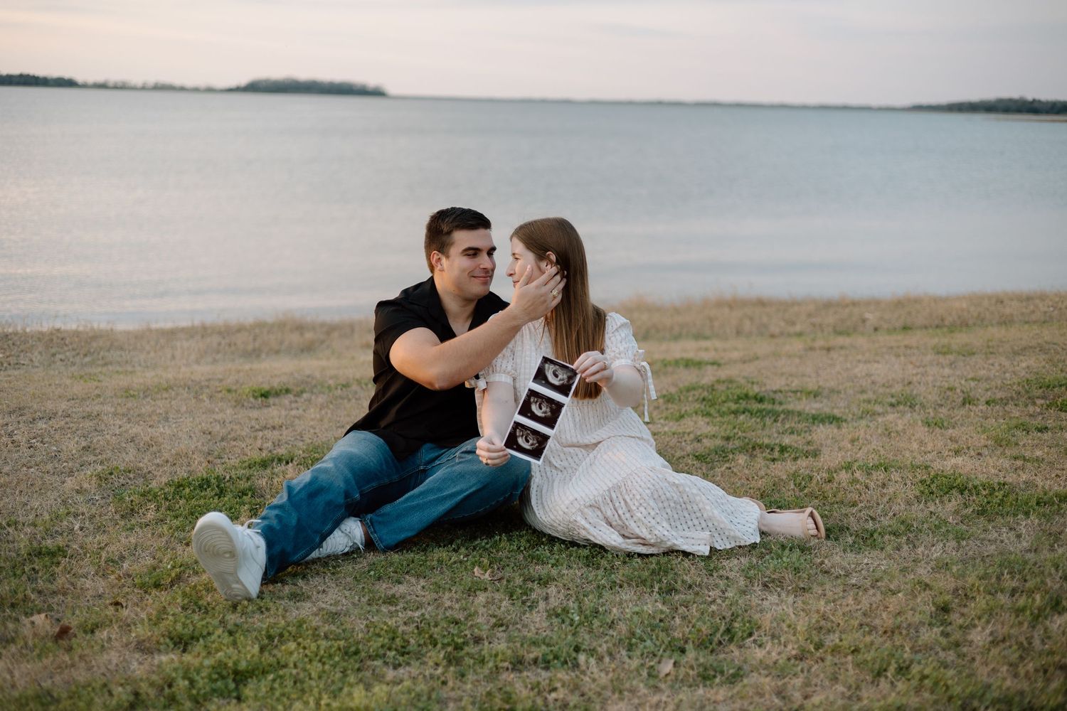Couple shares intimate moment sitting together on waterfront grass.