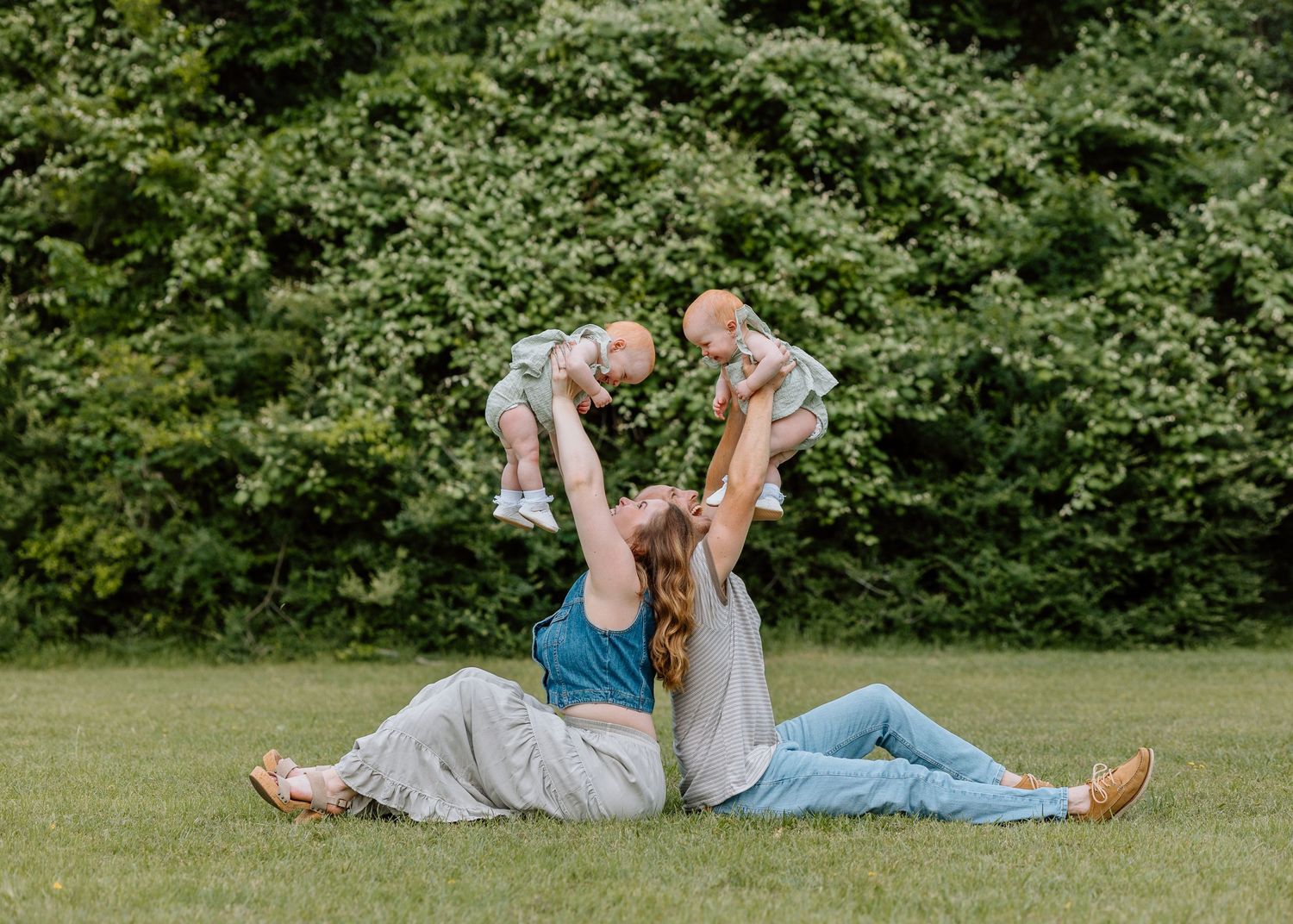 A family plays together on green grass in front of leafy bushes during a sunny summer day.