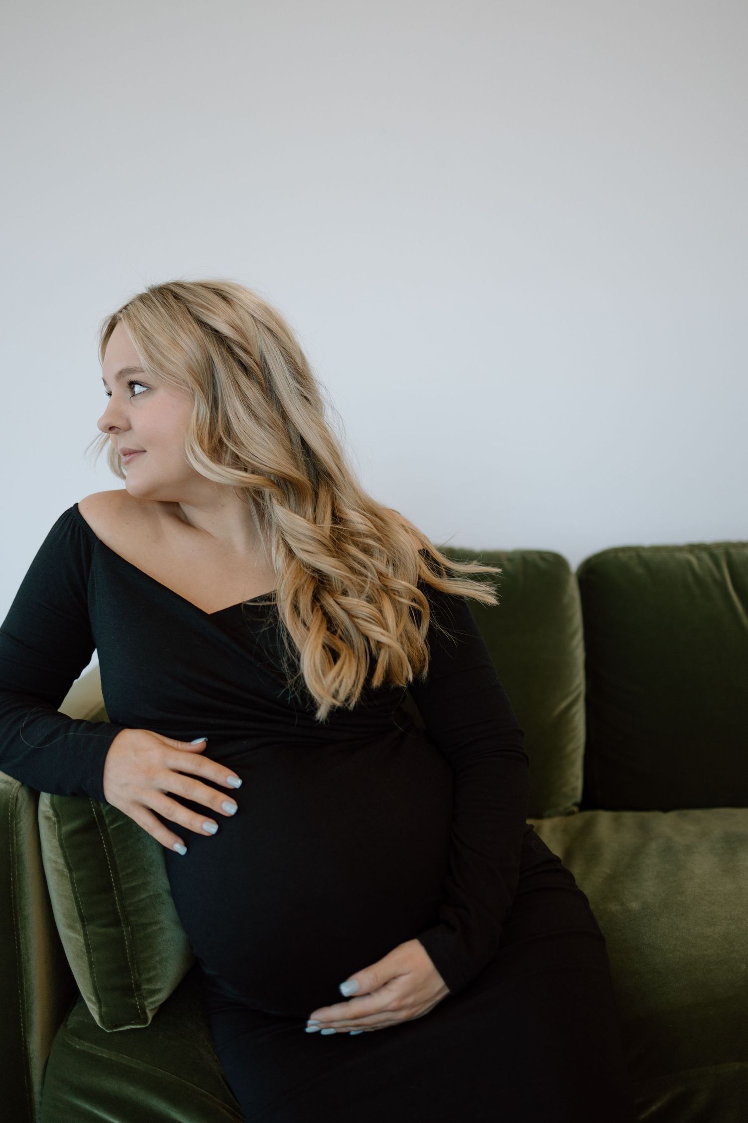 A pregnant woman in a black dress sits on a dark green velvet couch looking to the side.