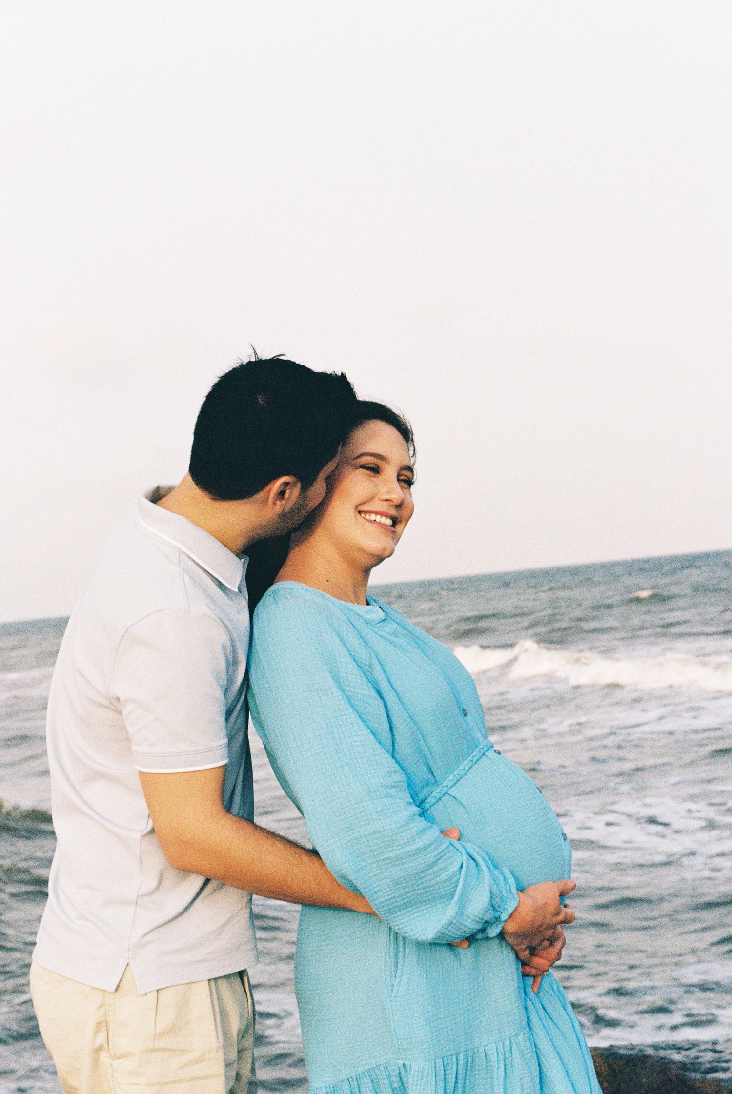 Couple embracing on beach as waves crash behind them during maternity photoshoot.