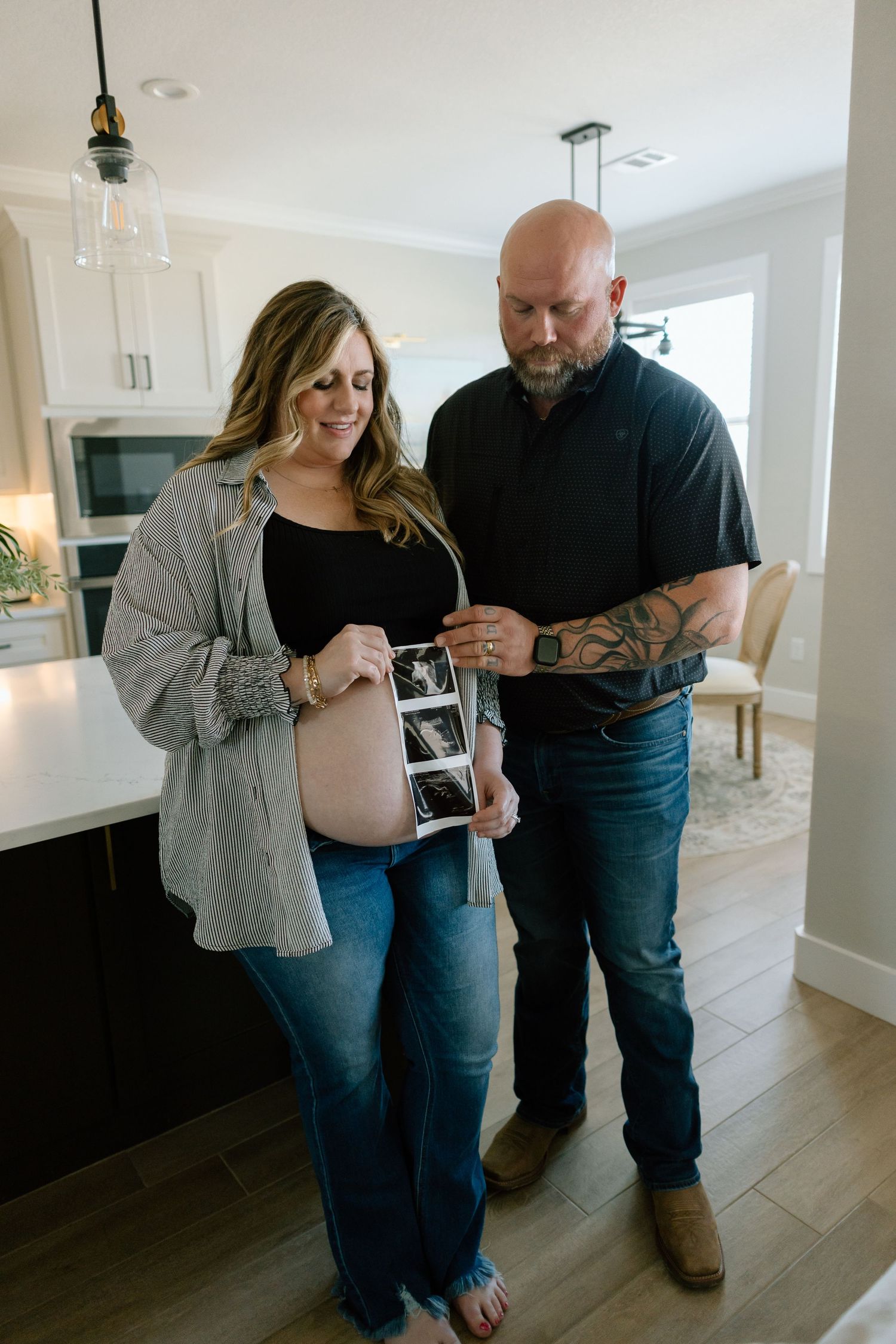 Couple in casual attire looks lovingly at ultrasound photos while standing in modern kitchen.