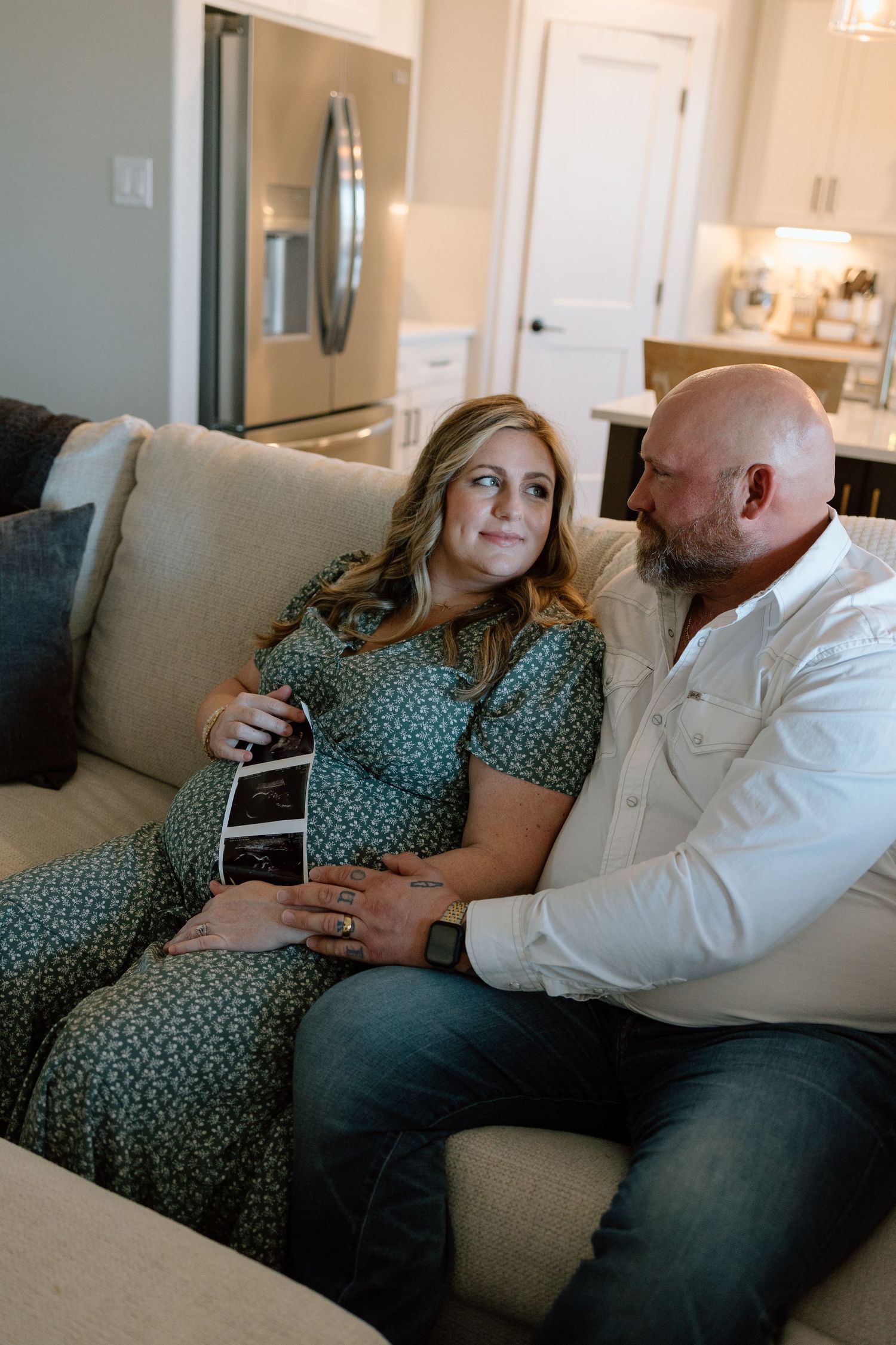A couple sits on a beige couch looking at an ultrasound photo together in a modern living room.