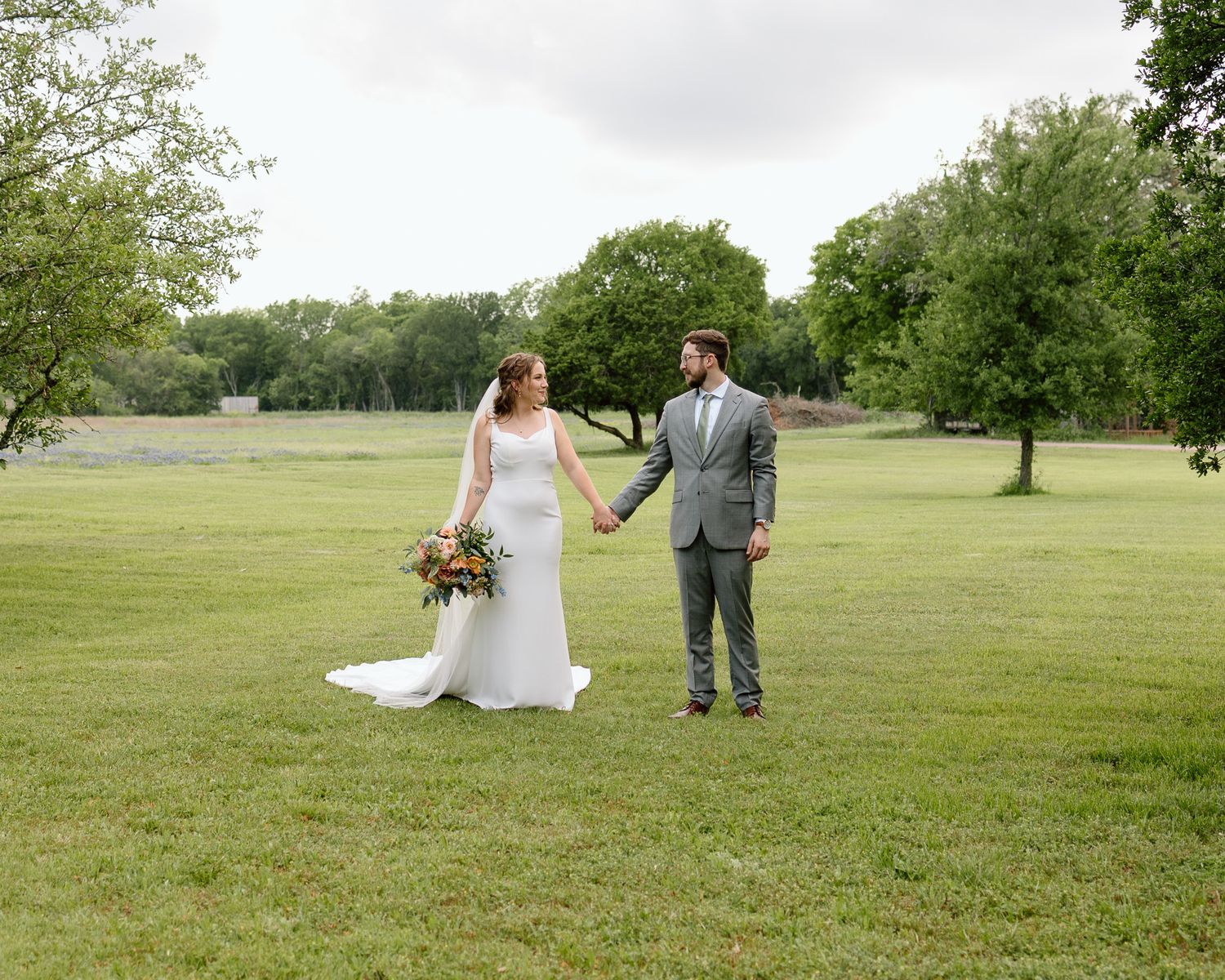 Wedding couple stands together on green lawn with trees and water feature in background.