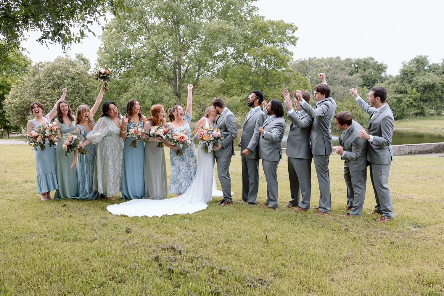 Wedding party in blue dresses and grey suits poses together on grassy lawn.