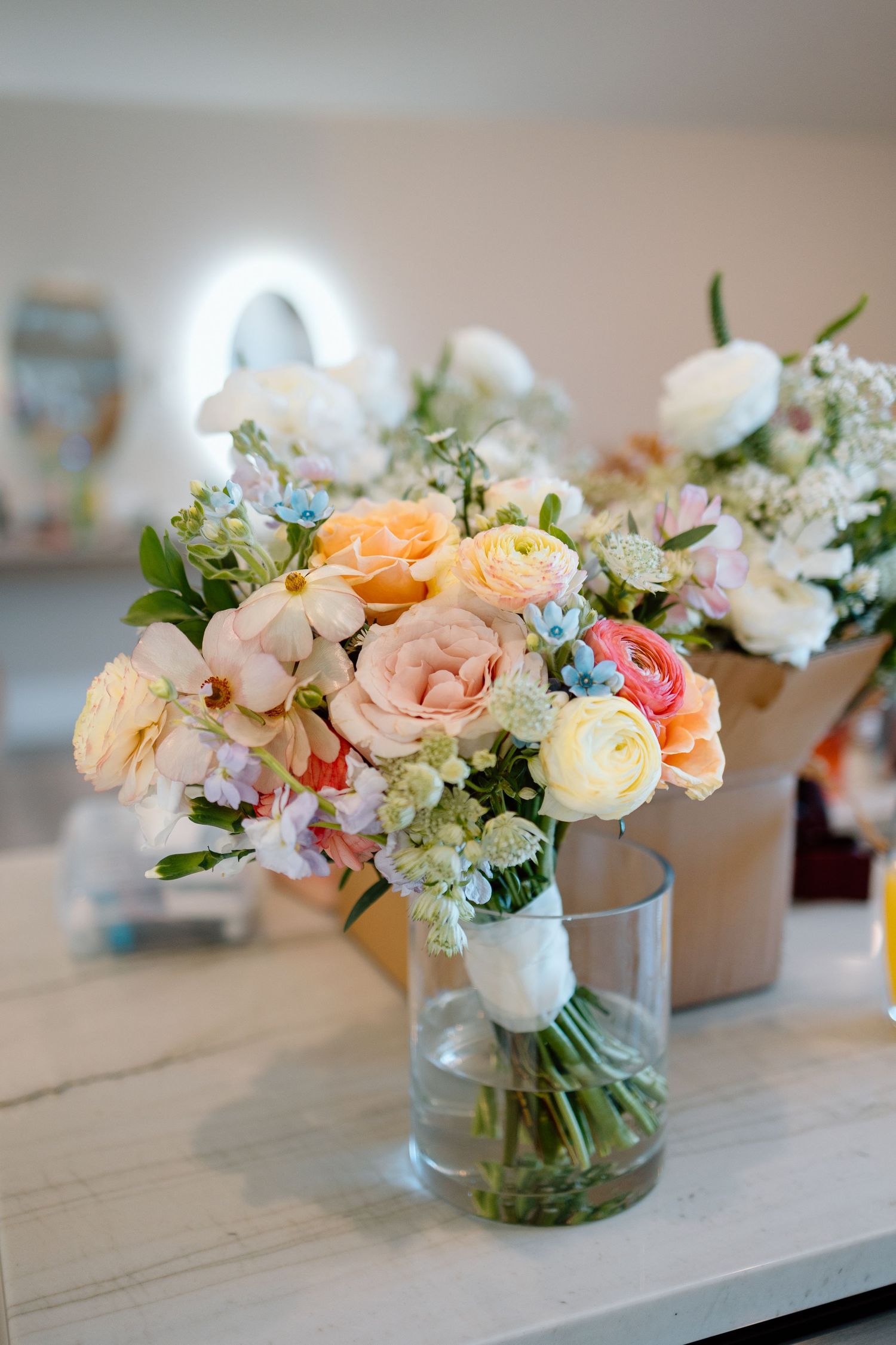 Pastel wedding bouquet with peach roses, white flowers and blue accents in glass vase.