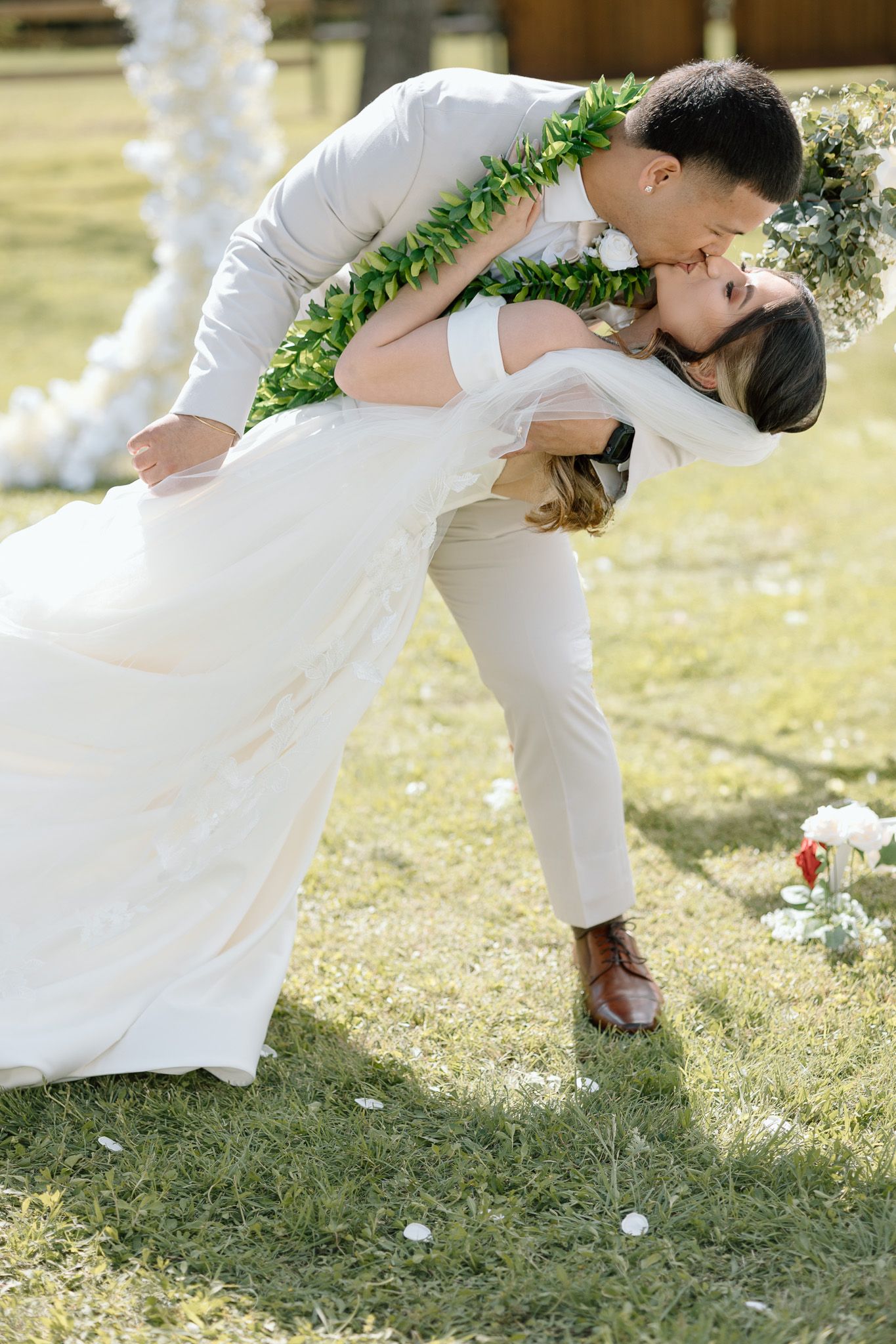 A romantic dip kiss between newlyweds wearing traditional white wedding attire and green lei garlands.