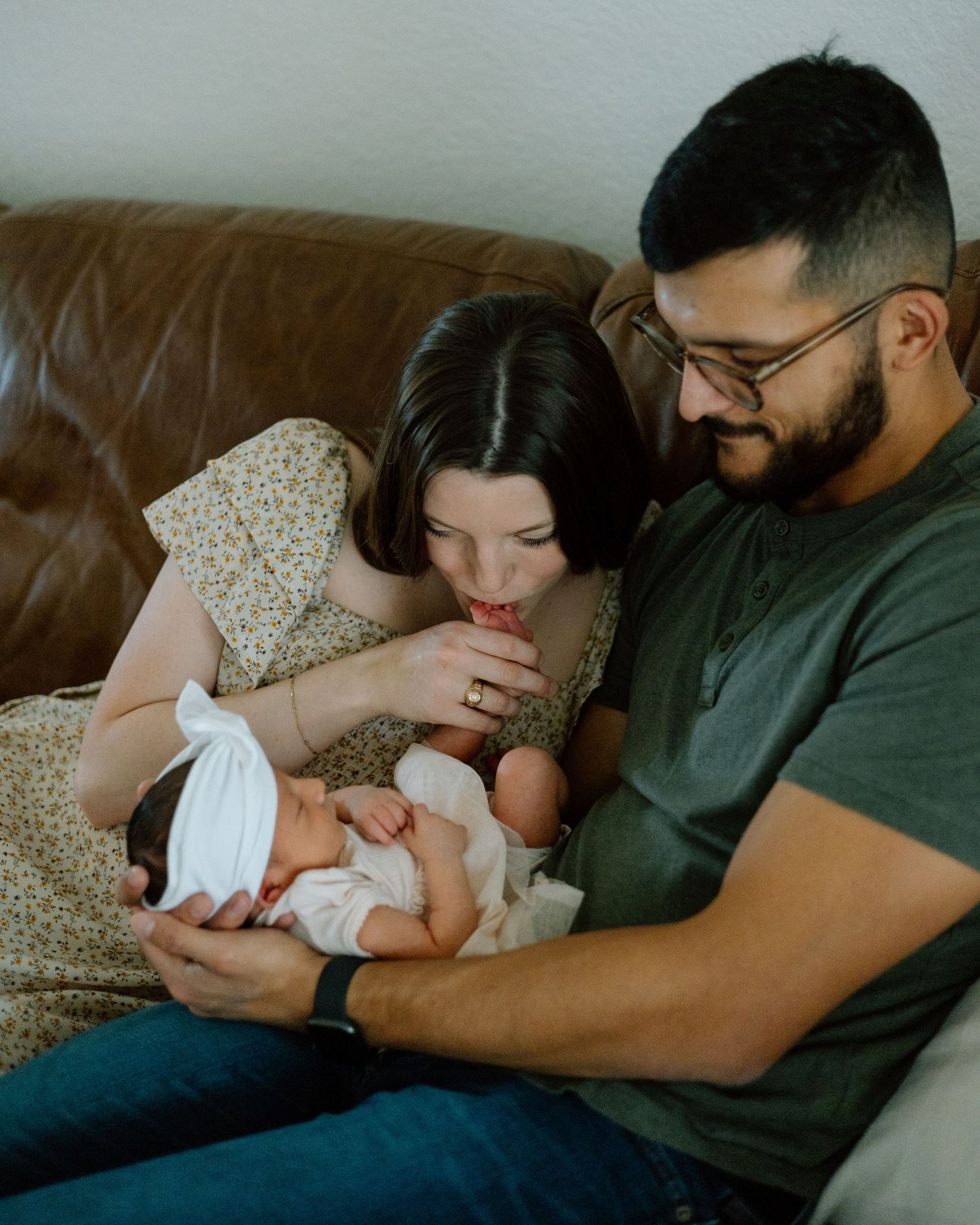 Family shares a tender moment on leather couch while holding their swaddled newborn.