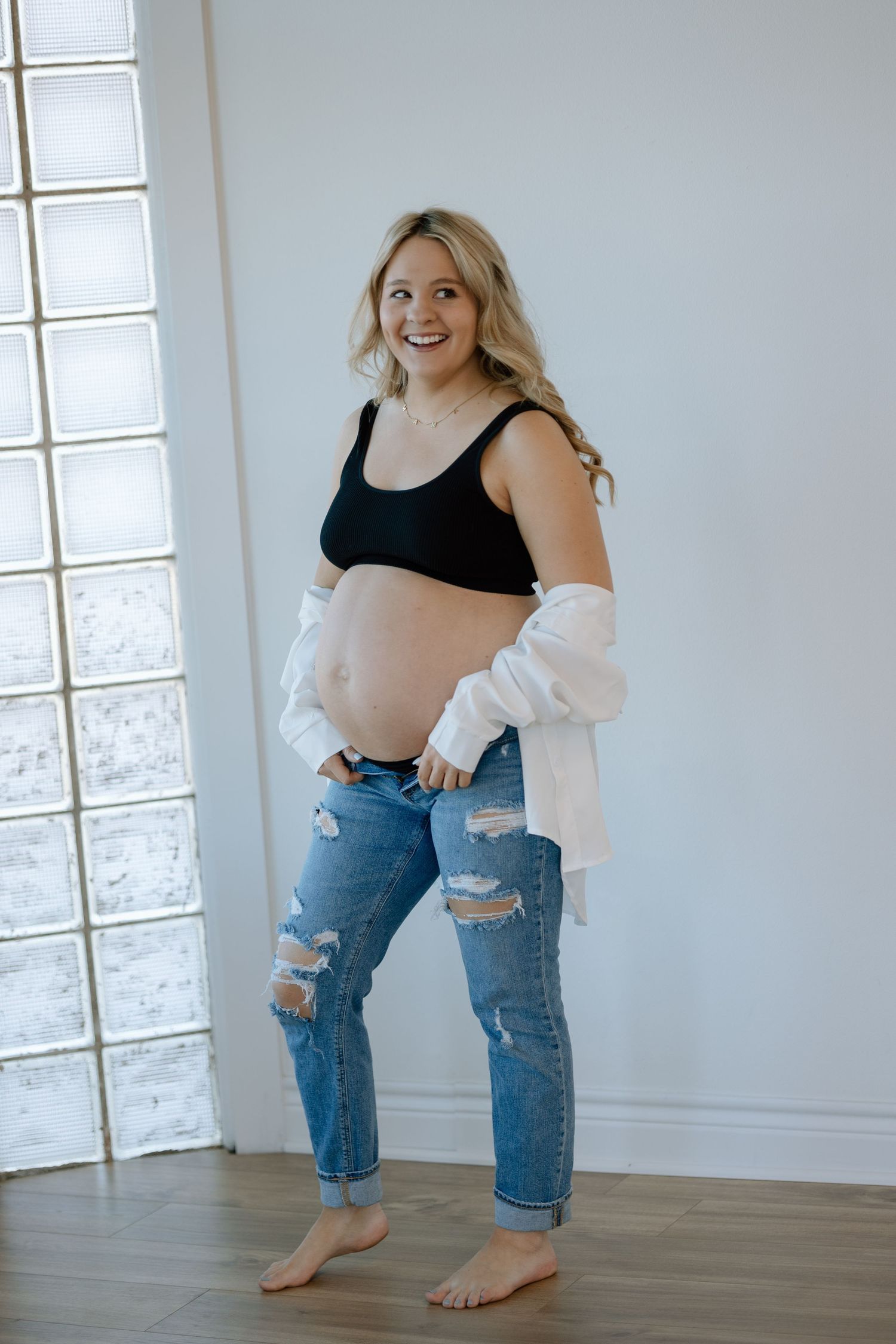 Pregnant woman in ripped jeans and black top poses barefoot against white wall with glass brick window.