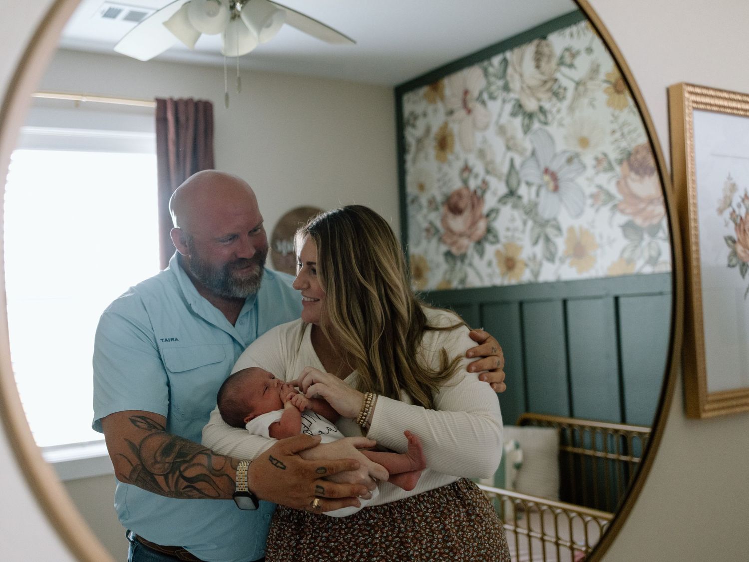 Couple in nursery room holding newborn baby near window with floral wallpaper and board and batten wall details.