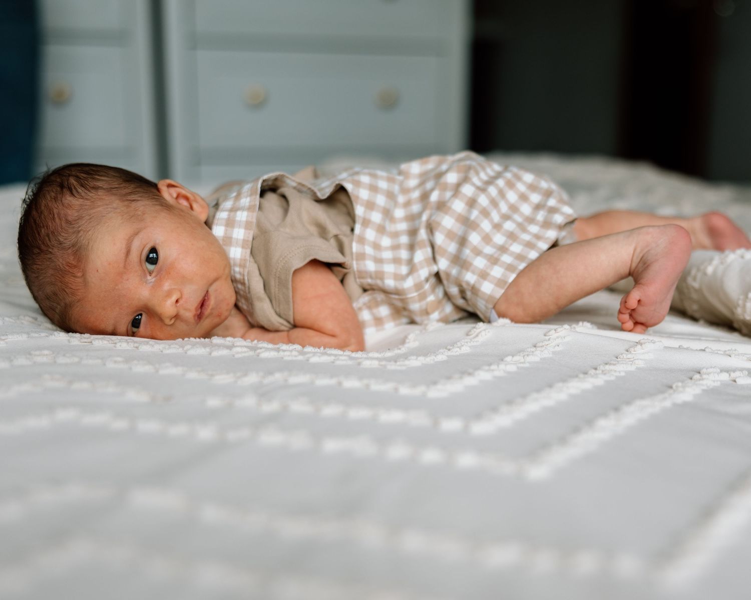 Newborn baby in checkered outfit lying on white textured blanket, looking at camera with alert eyes.