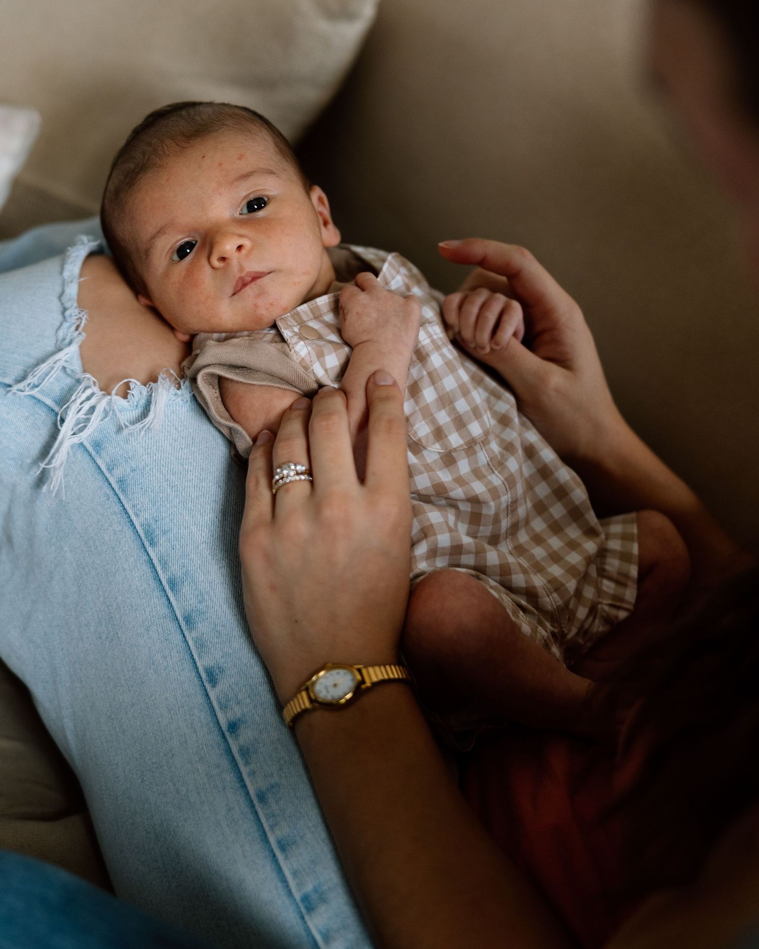 Newborn baby in gingham onesie cradled in mother's arms, wearing gold watch and diamond ring.