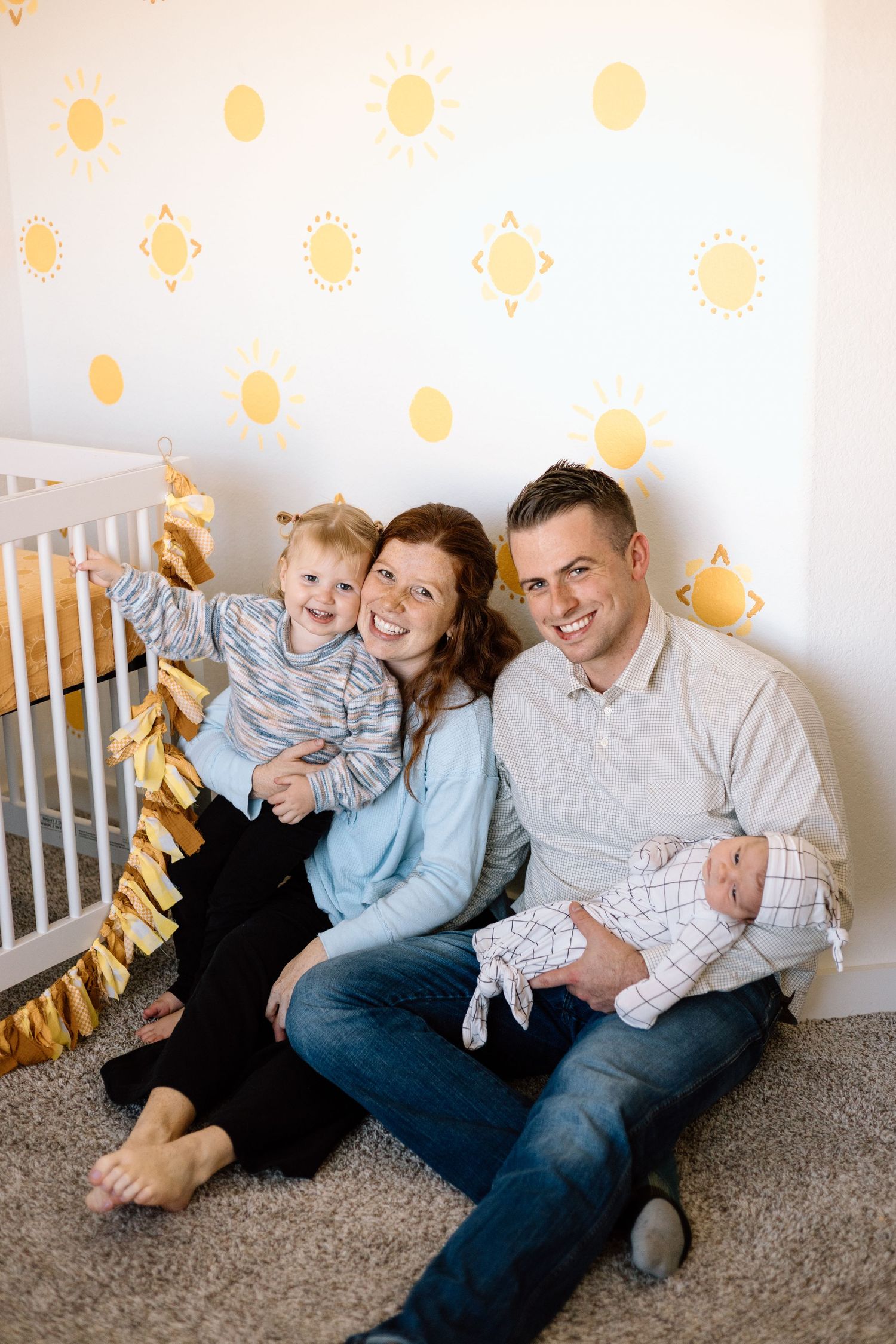 Happy family of four sitting on nursery floor with sun-themed wall decals, parents smiling with toddler and newborn baby.