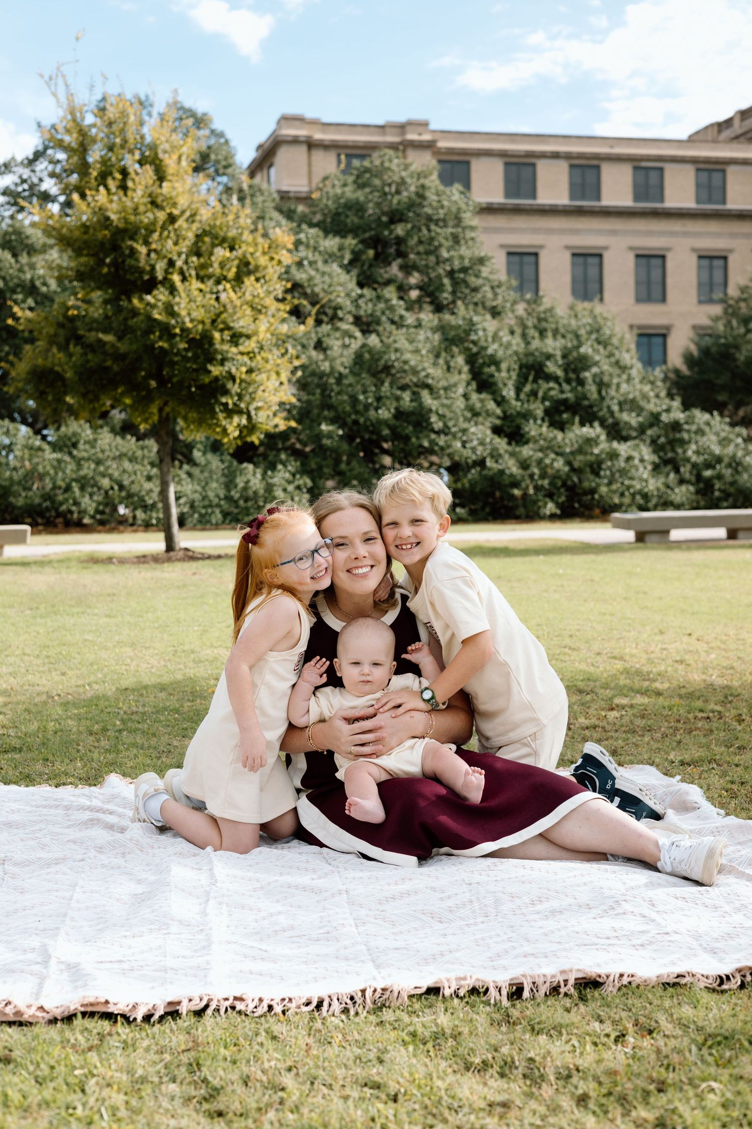 A mother sits on a blanket in a park, smiling with three children, including a baby, on a sunny day.