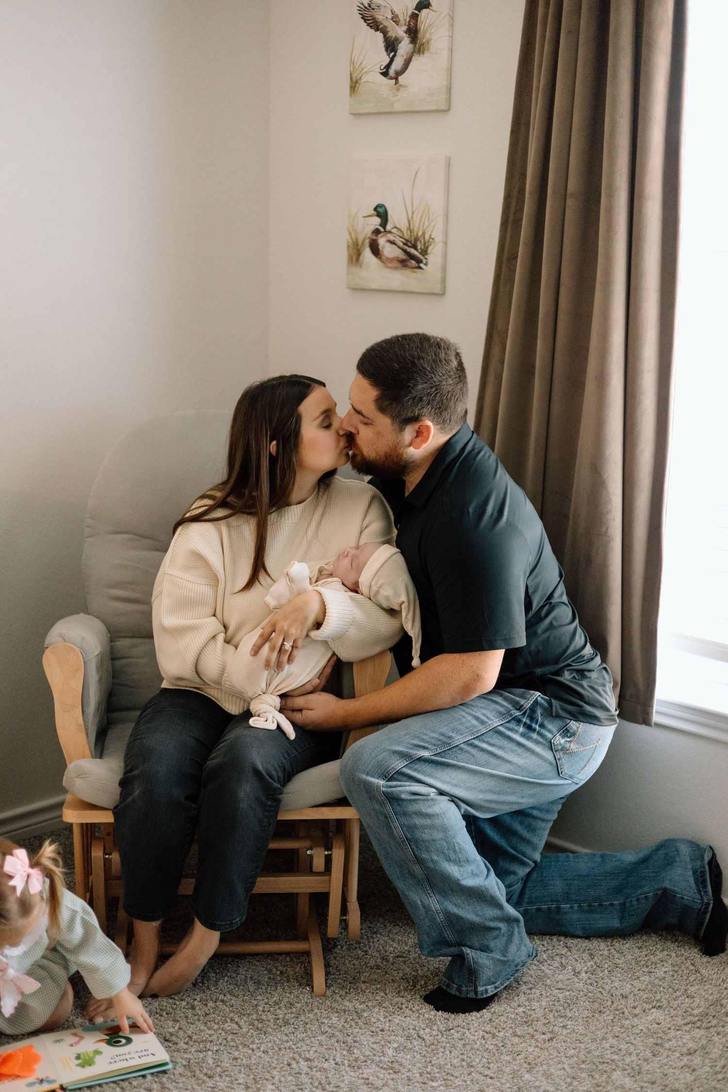 New parents kissing while mother holds newborn baby in nursery chair with duck artwork on wall.