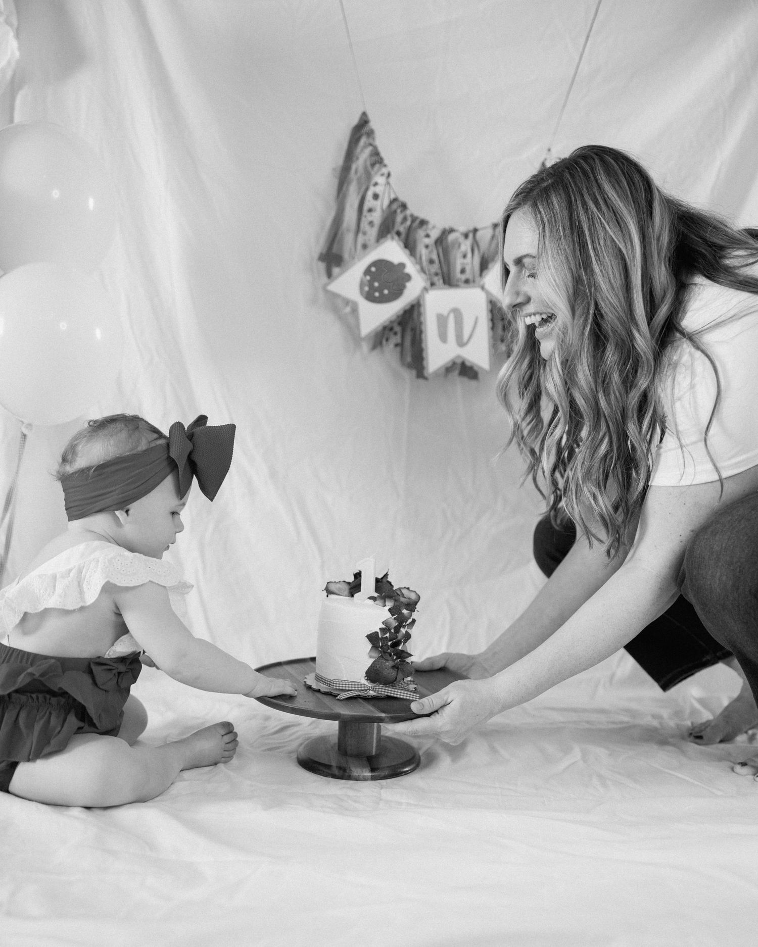 Black and white photo of a smiling mother presenting a small cake to her baby during a first birthday cake smash photoshoot.