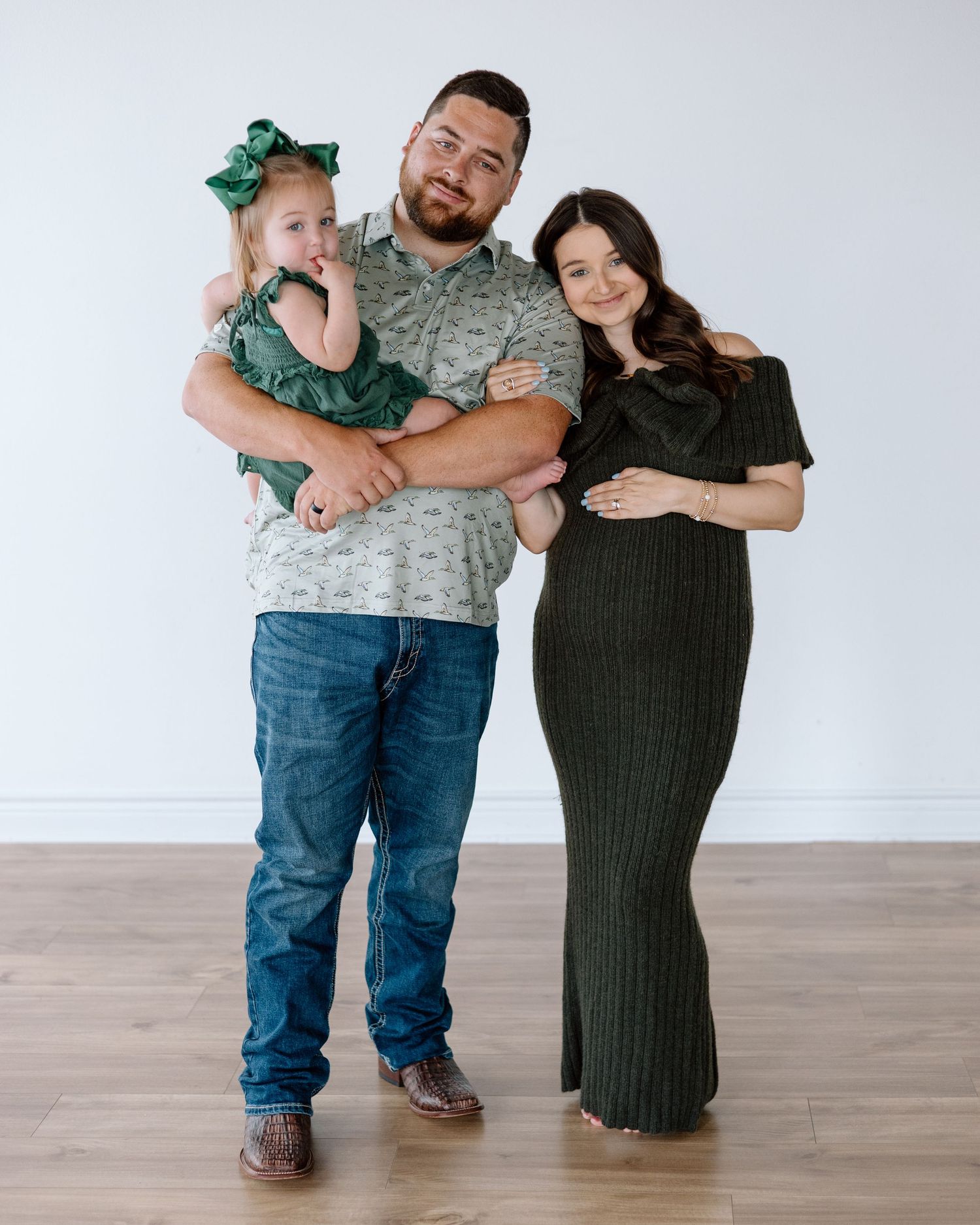 A family of three poses together, man holding toddler in green dress, pregnant woman in dark olive maxi dress.