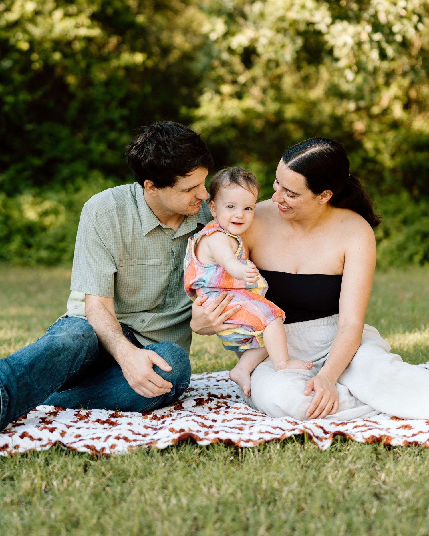 A happy young family sits on a blanket in a park, smiling at their baby girl in a colorful dress.